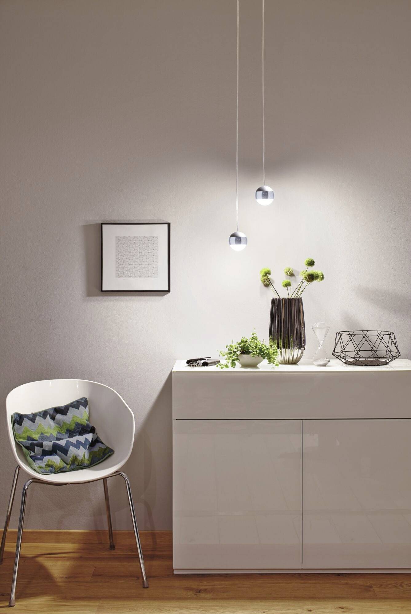 Ein minimalistisches Wohnzimmer mit weißem Sideboard und moderner Dekoration. Hängelampen beleuchten frische Pflanzen und geometrische Vasen.