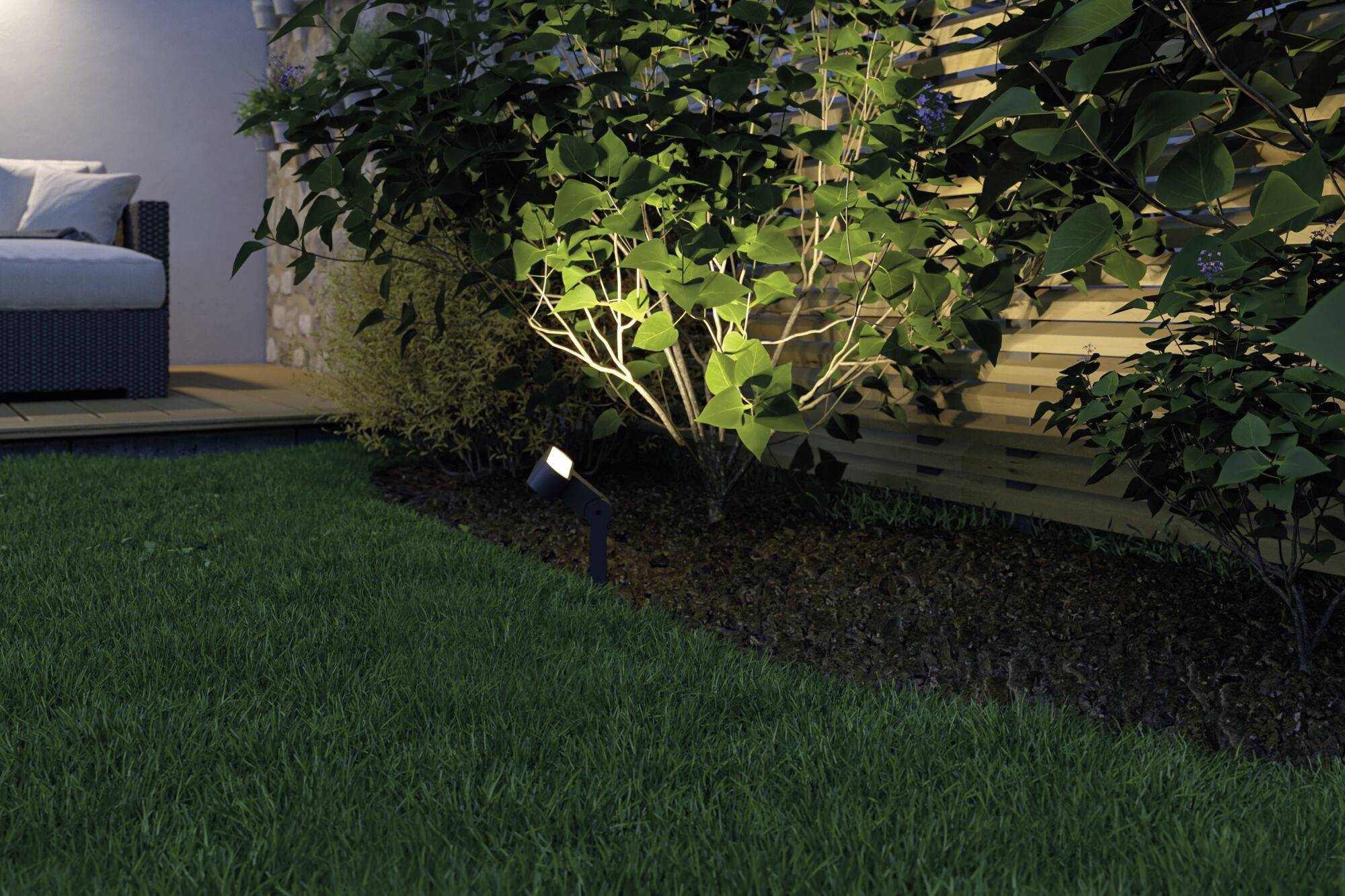 Ein Garten mit Rasen, beleuchtet von einem kleinen warmen Spotlight, das auf einen Strauch gerichtet ist, nahe einer Terrasse.