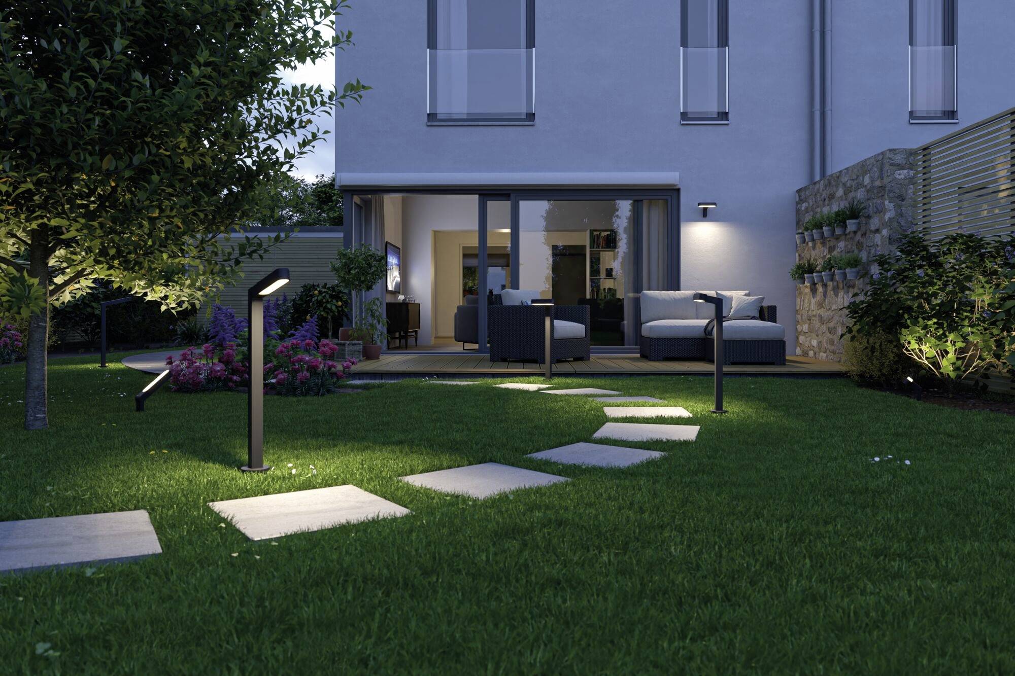 Ein Garten bei Nacht, beleuchtet durch Weglampen, führt zu einer Terrasse. Auf der Terrasse sind Sofas und ein Tisch vor einem modernen Haus.