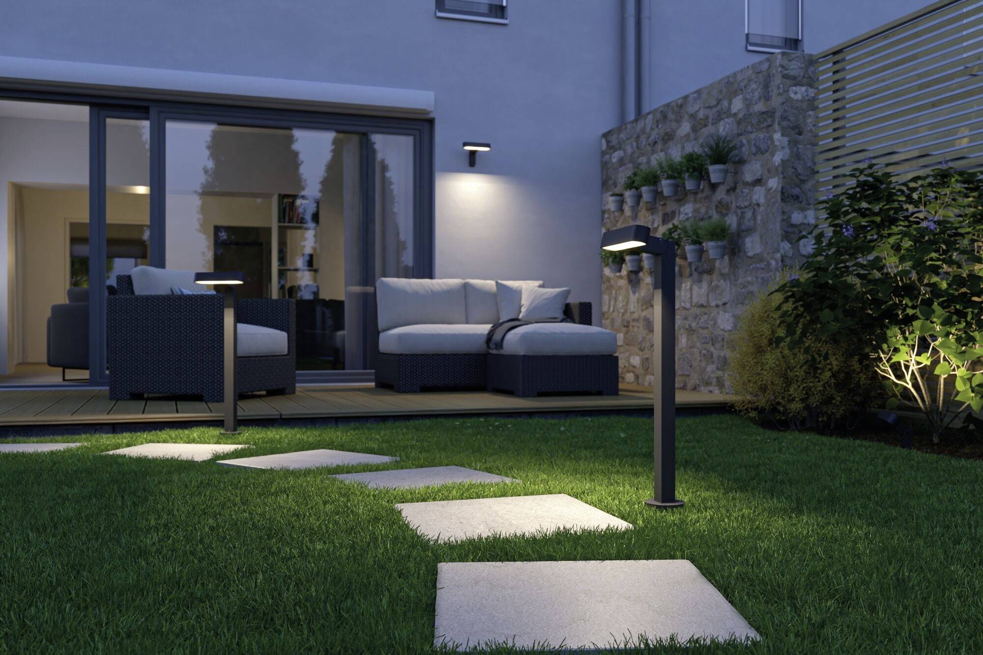 Ein moderner Garten bei Abenddämmerung: beleuchtete Gehwegplatten führen zu einer Terrasse mit weißen Sofas, umgeben von Pflanzen.