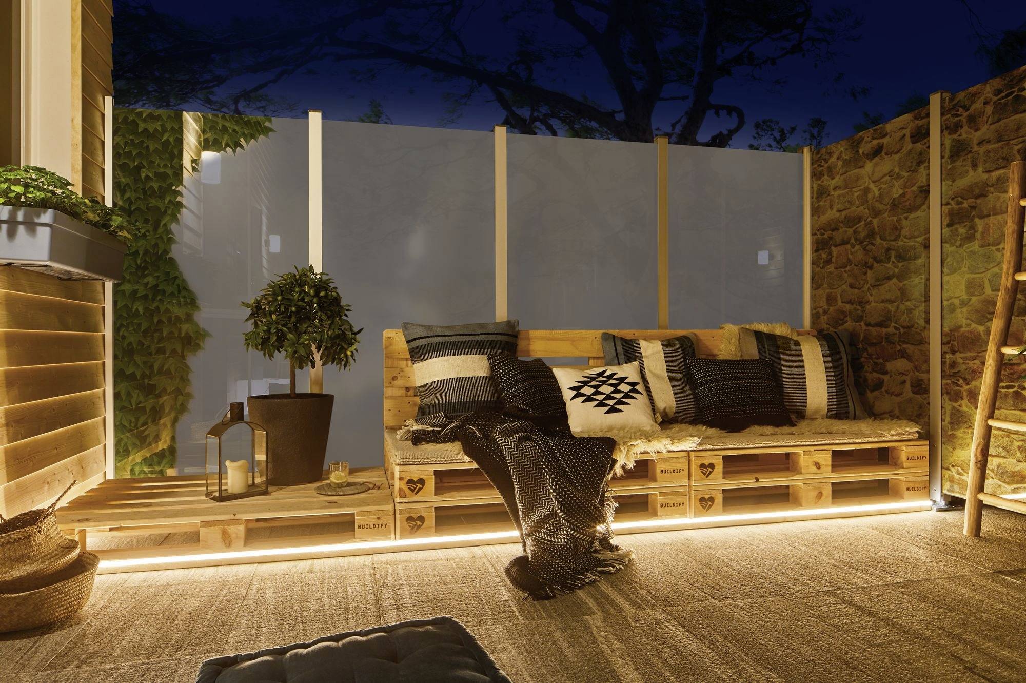 Gemütliche Terrasse bei Nacht mit Holzpaletten-Sofa, Kissen und Decken; weiche Beleuchtung erzeugt eine einladende Atmosphäre.