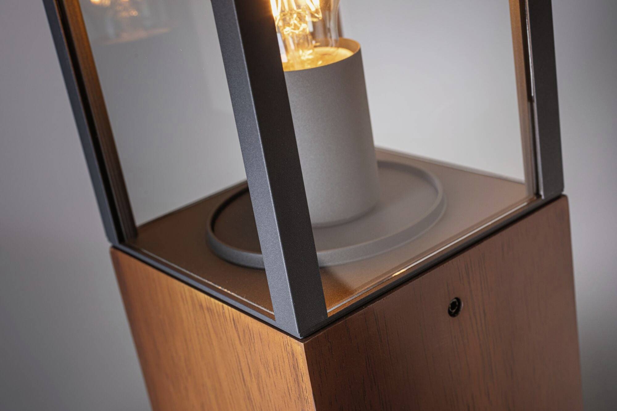 Eine moderne, kubische Tischleuchte aus Holz und Glas mit sichtbarem Leuchtmittel, das warmes Licht abstrahlt.