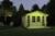 Ein beleuchtetes Gartenhaus in einer abendlichen Umgebung, umgeben von Bäumen. Vor dem Haus stehen ein Tisch und zwei Stühle.