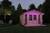 Ein kleines Holzhaus mit rosa Beleuchtung steht im Garten. Daneben sind Gartenmöbel und ein beleuchteter Baum. Abendszene im Freien.