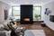 Ein modernes Wohnzimmer mit Kamin in der Mitte, umgeben von Pflanzen und Holz. Eine Couch links und ein Fernseher an der Wand rechts.