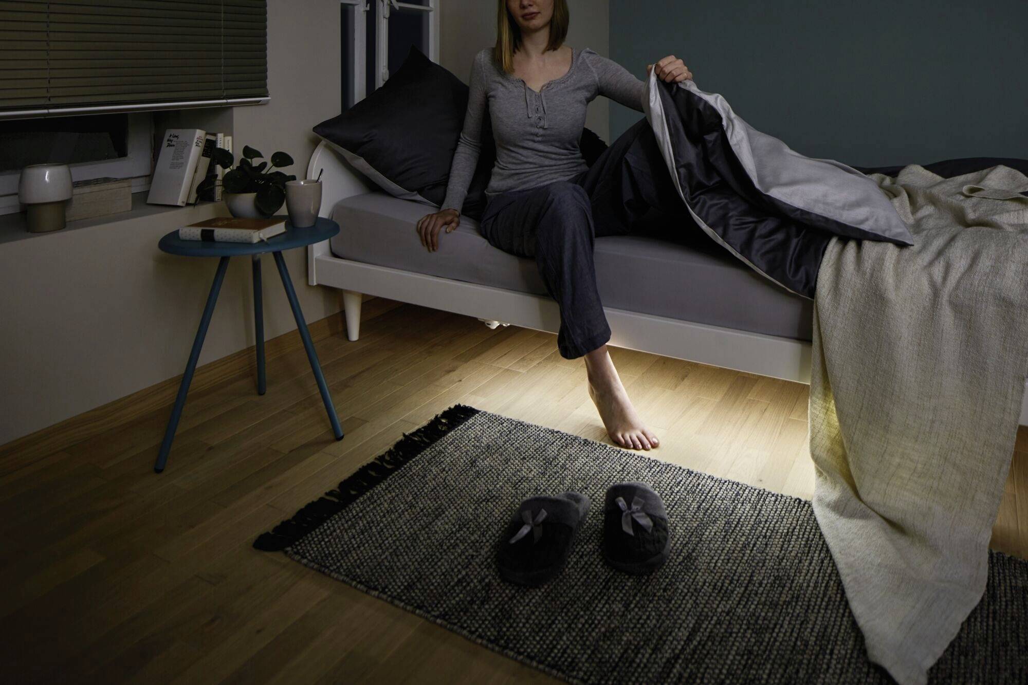 Eine Frau sitzt auf einem Bett in einem dunklen Raum. Ein Licht leuchtet unter dem Bett hervor, Sessel mit Hausschuhen auf einem Teppich.
