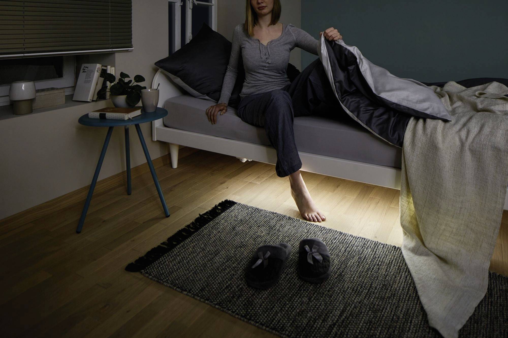 Eine Person sitzt auf einem Bett und hält die Bettdecke hoch. Auf dem Boden sind Hausschuhe vor einem Teppich zu sehen.
