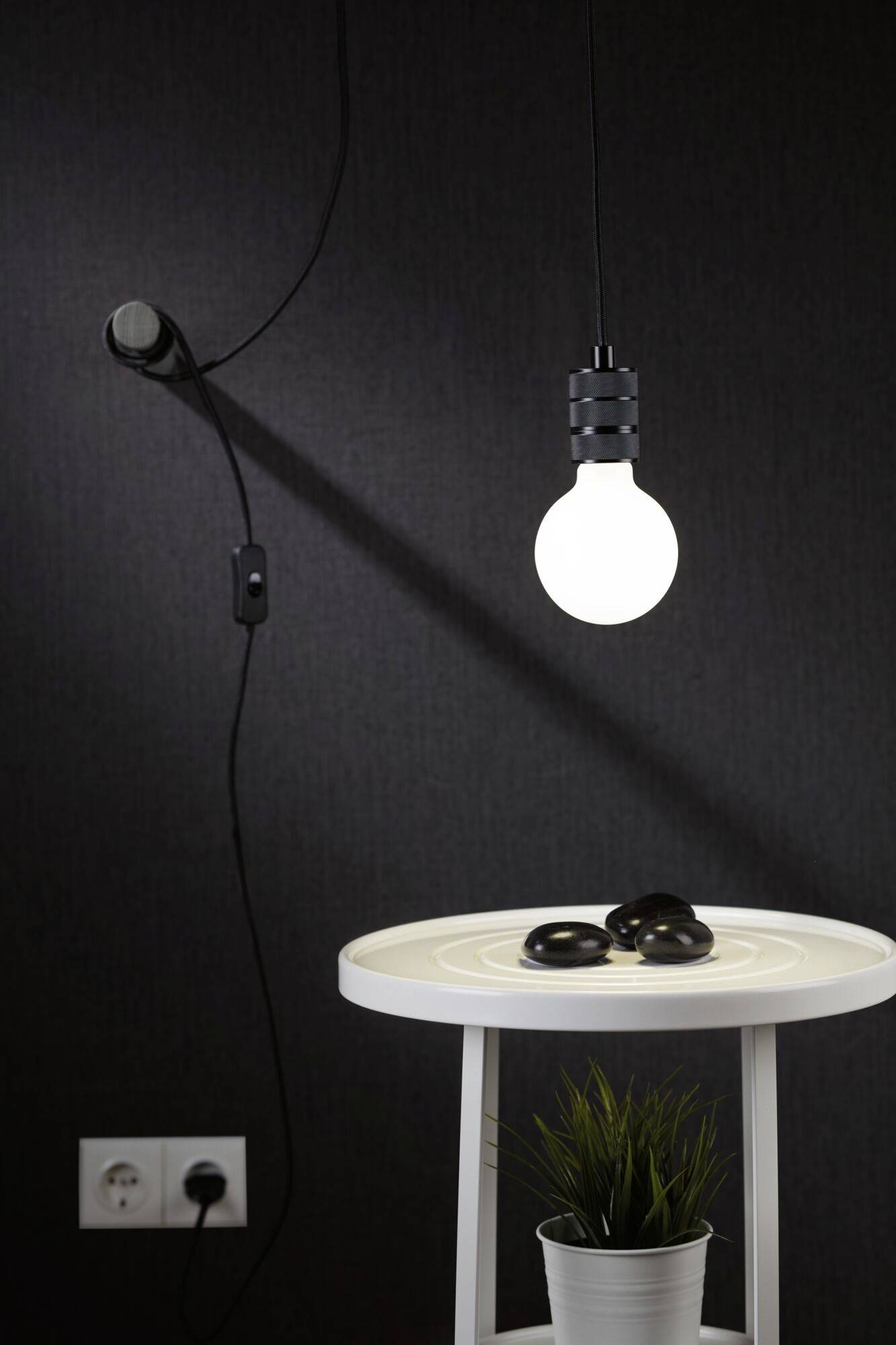 Eine weiße Tischlampe mit schwarzem Kabel und Schalter, eine leuchtende Glühbirne und ein kleiner Blumentopf stehen auf einem Tisch.
