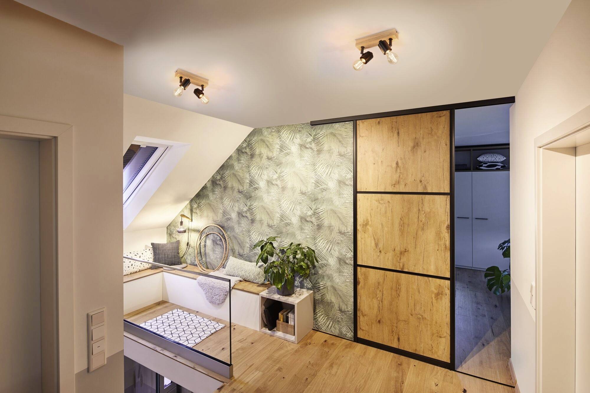Ein modernes, helles Interieur eines Dachzimmers mit Holz- und Pflanzenmotiven. Ein Sitzbereich mit Kissen auf der linken Seite.