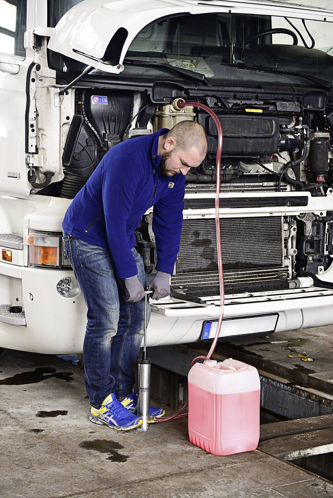 Mann in blauer Jacke und Jeans füllt Flüssigkeit mit einer Pumpe aus einem roten Kanister in ein Fahrzeug mit offenem Motorraum.