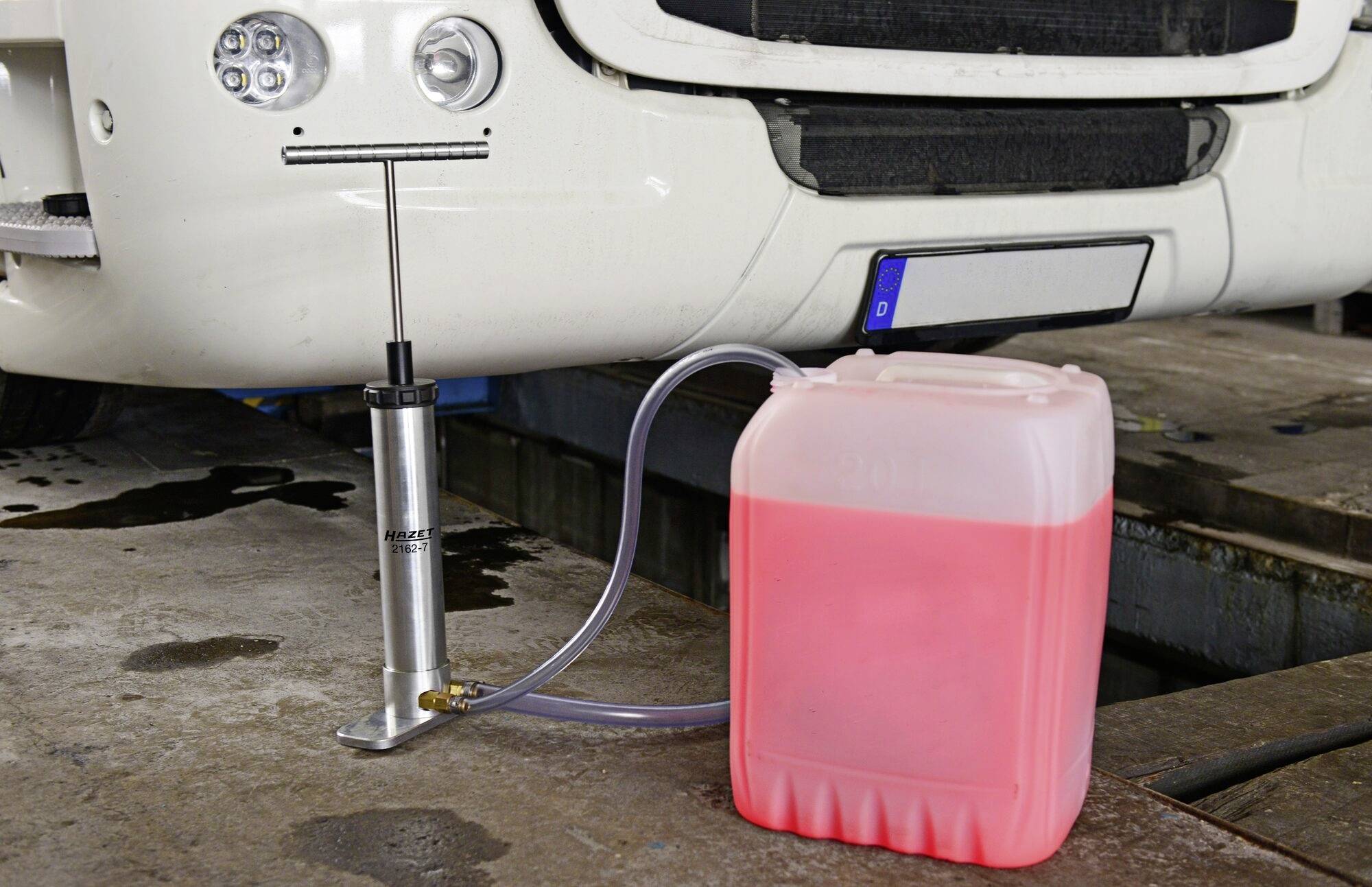 Ein weißes Fahrzeug hebt sich mit einem hydraulischen Wagenheber, daneben steht ein Behälter mit roter Flüssigkeit und einem Schlauch.