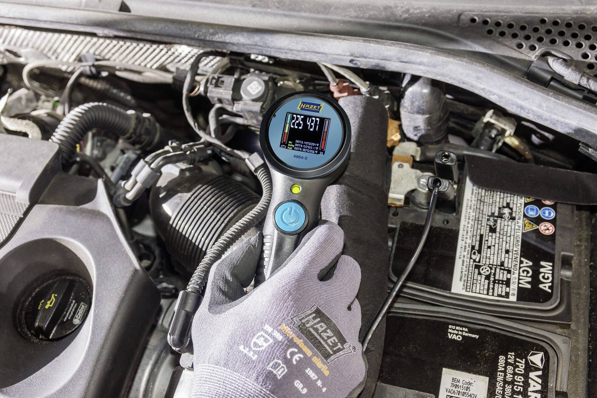 Eine Person misst mit einem digitalen Gerät unter einer geöffneten Autokohle den Zustand einer Autobatterie, die 12,85 Volt anzeigt.