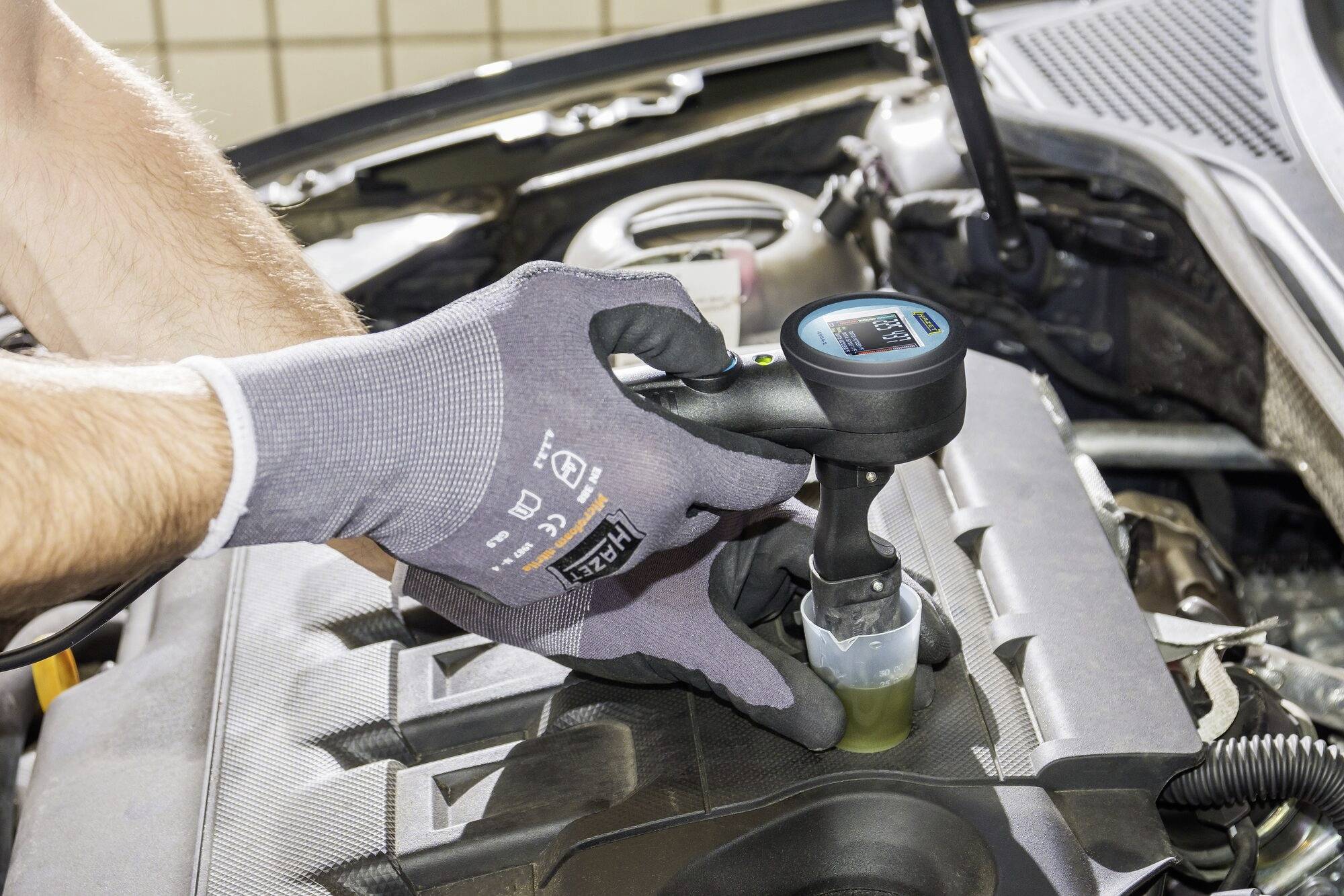 Eine Person testet Kühlflüssigkeit im Motorraum eines Autos. Sie trägt Handschuhe und benutzt ein digitales Messgerät.
