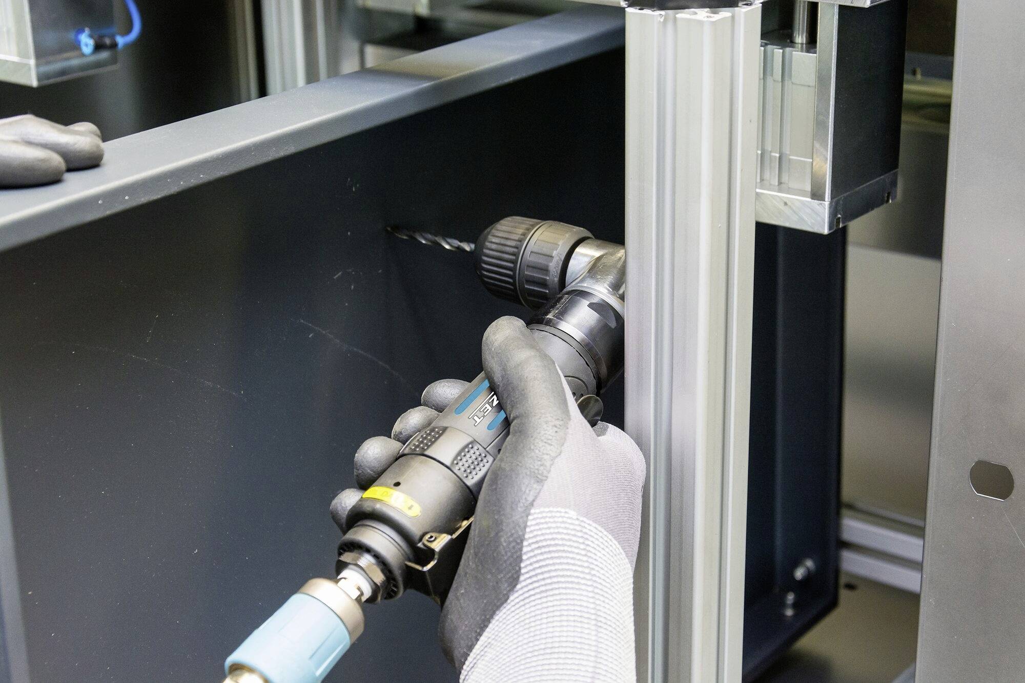 Eine Person mit Handschuhen verwendet einen elektrischen Bohrer, um ein Loch in eine Metallplatte in einer Werkstattumgebung zu bohren.