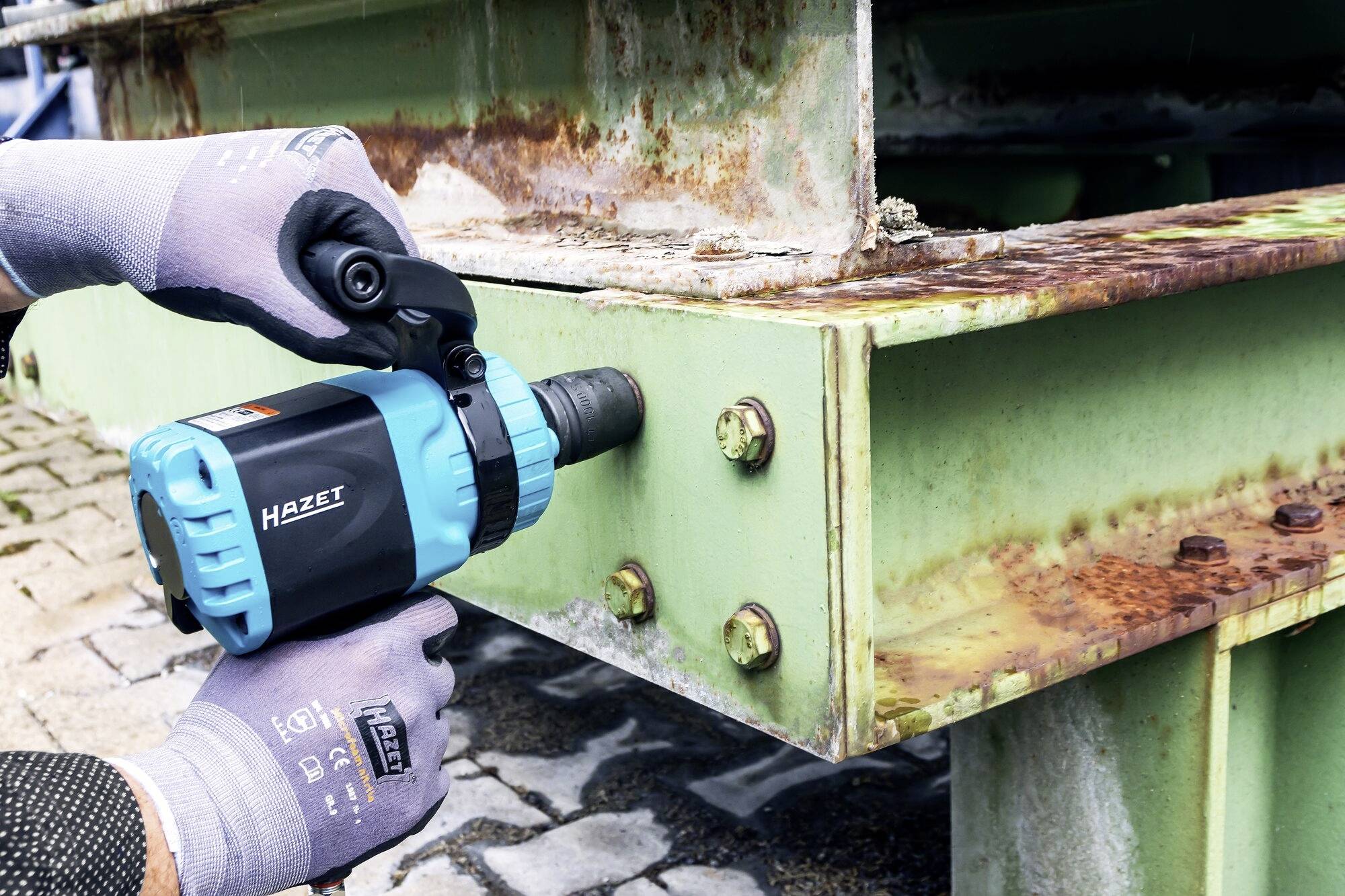 Eine Person zieht mit einem blauen, elektrischen Schlagschrauber Schrauben an einem rostigen Metallträger fest.