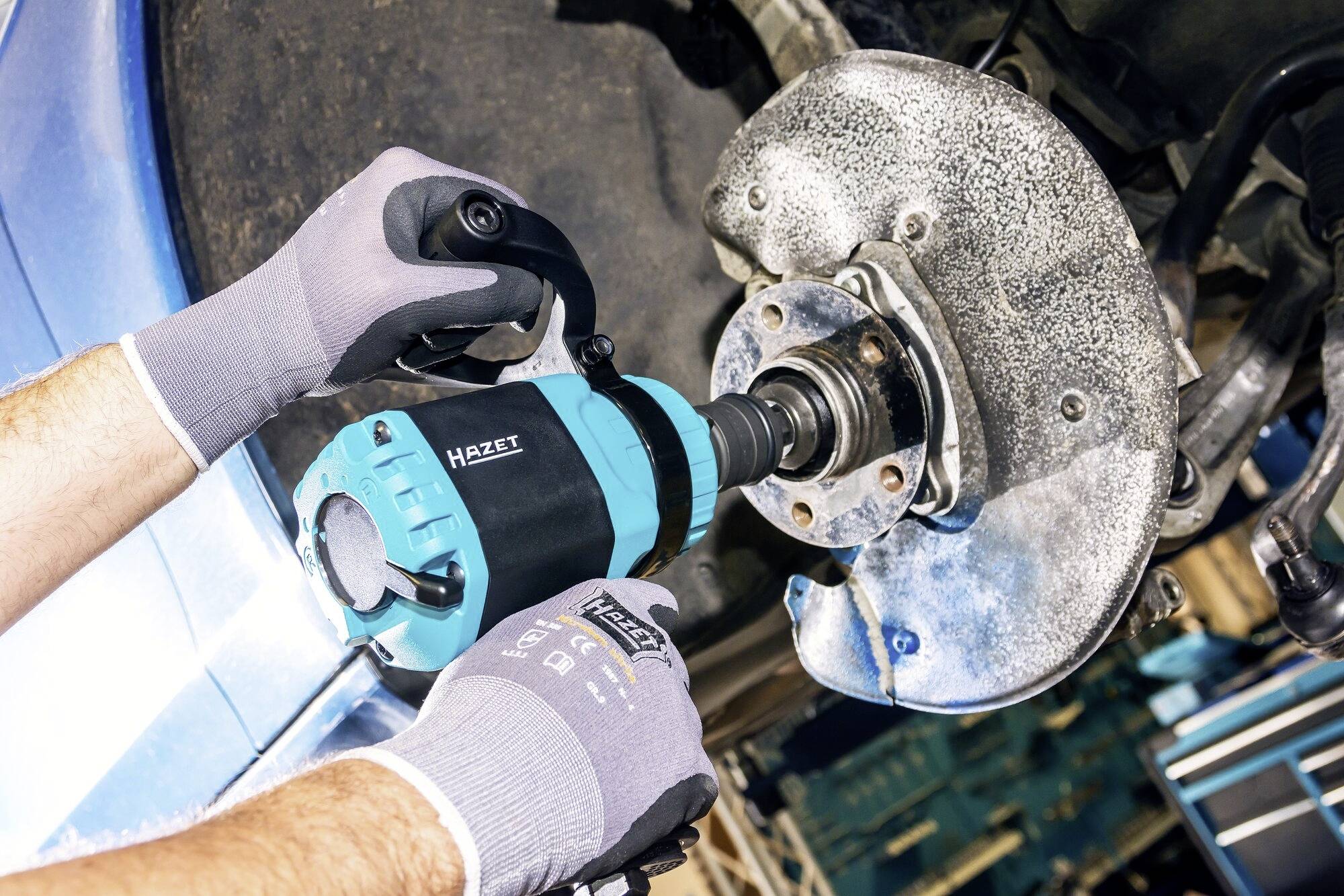 'Hände mit Arbeitshandschuhen benutzen einen Schlagschrauber der Marke Hazet, um eine Schraube an einem Autoteil zu lösen.'