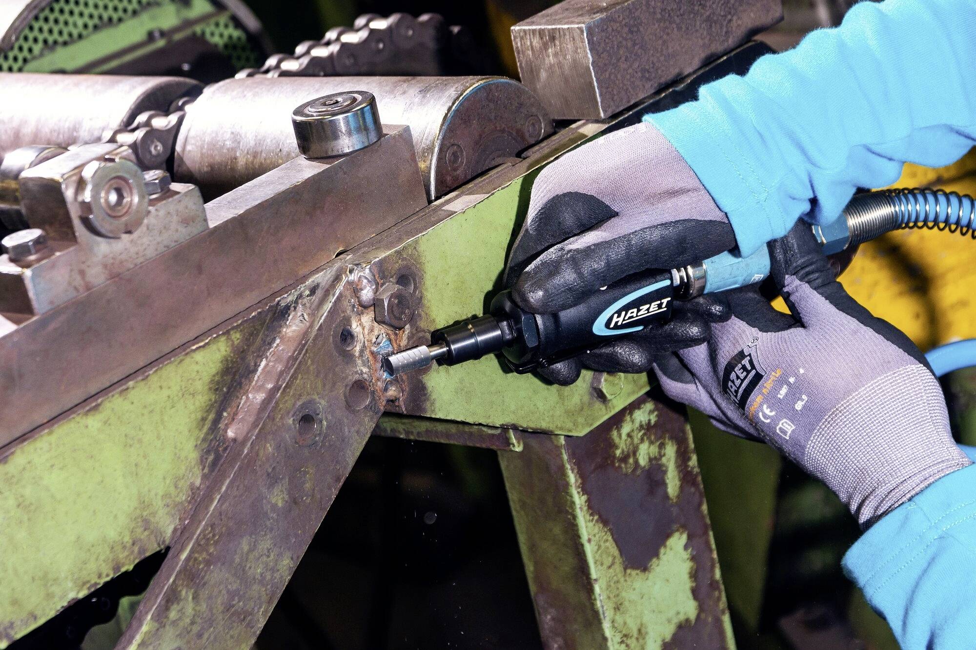 Eine Person in blauen Handschuhen arbeitet mit einem Druckluftschrauber an einer metallischen Rahmenkonstruktion in einer Werkstatt.