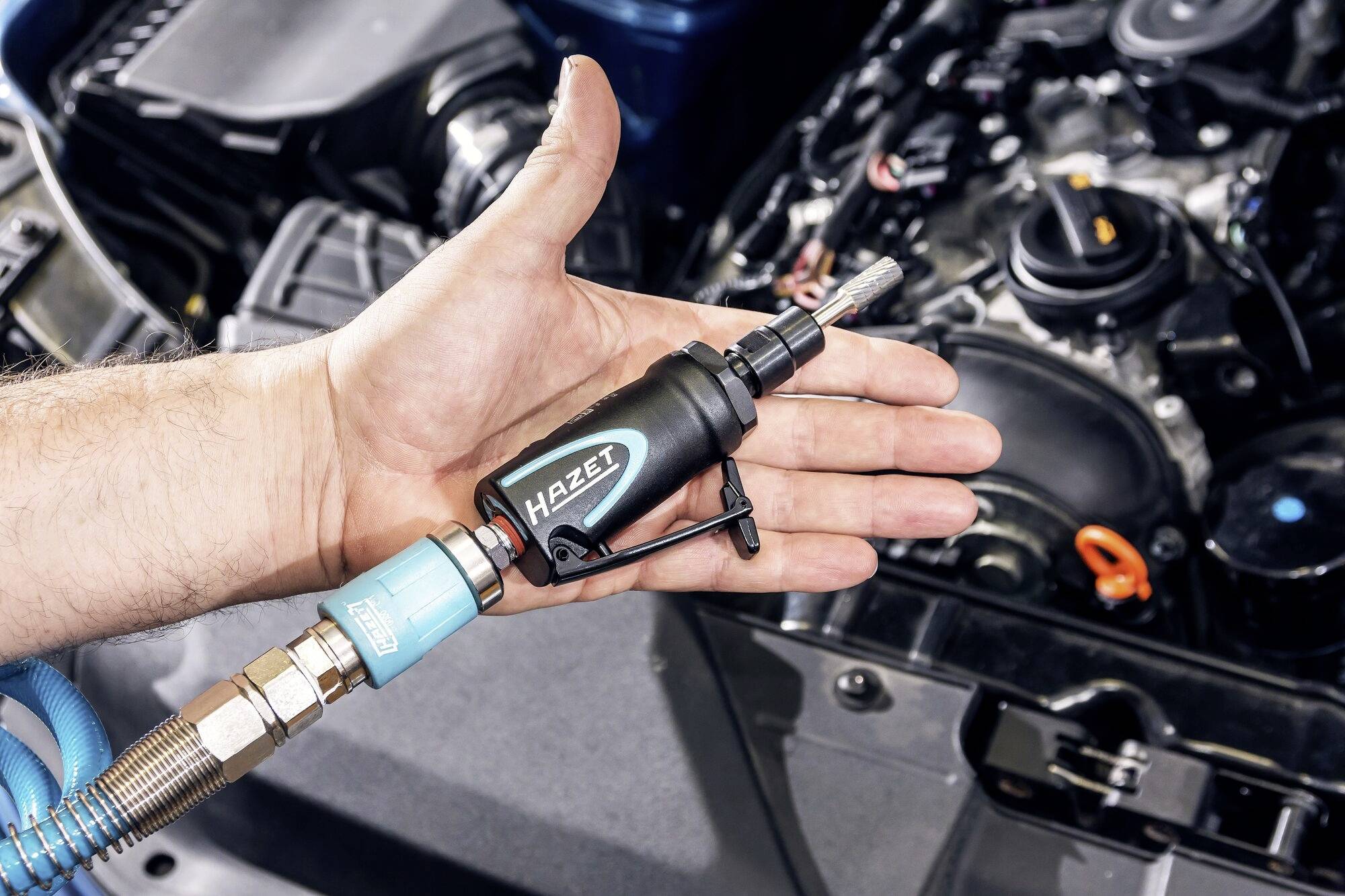 Eine Hand hält ein 'Hazet'-Werkzeug mit pneumatischem Anschluss vor einem offenen Motorraum eines Autos, bereit zur Benutzung.
