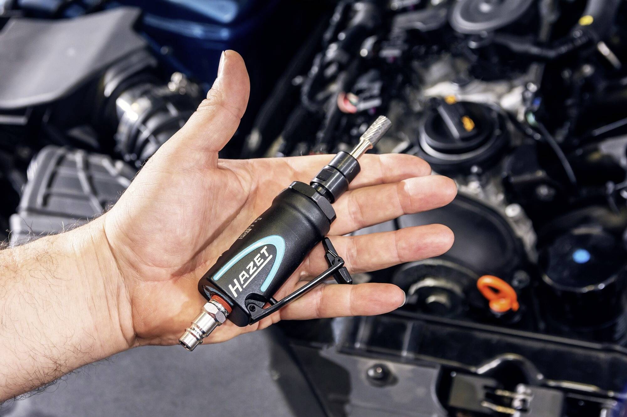 Eine Hand hält ein kleines pneumatisches Werkzeug mit der Aufschrift 'Hazet' vor einem geöffneten Automotor.