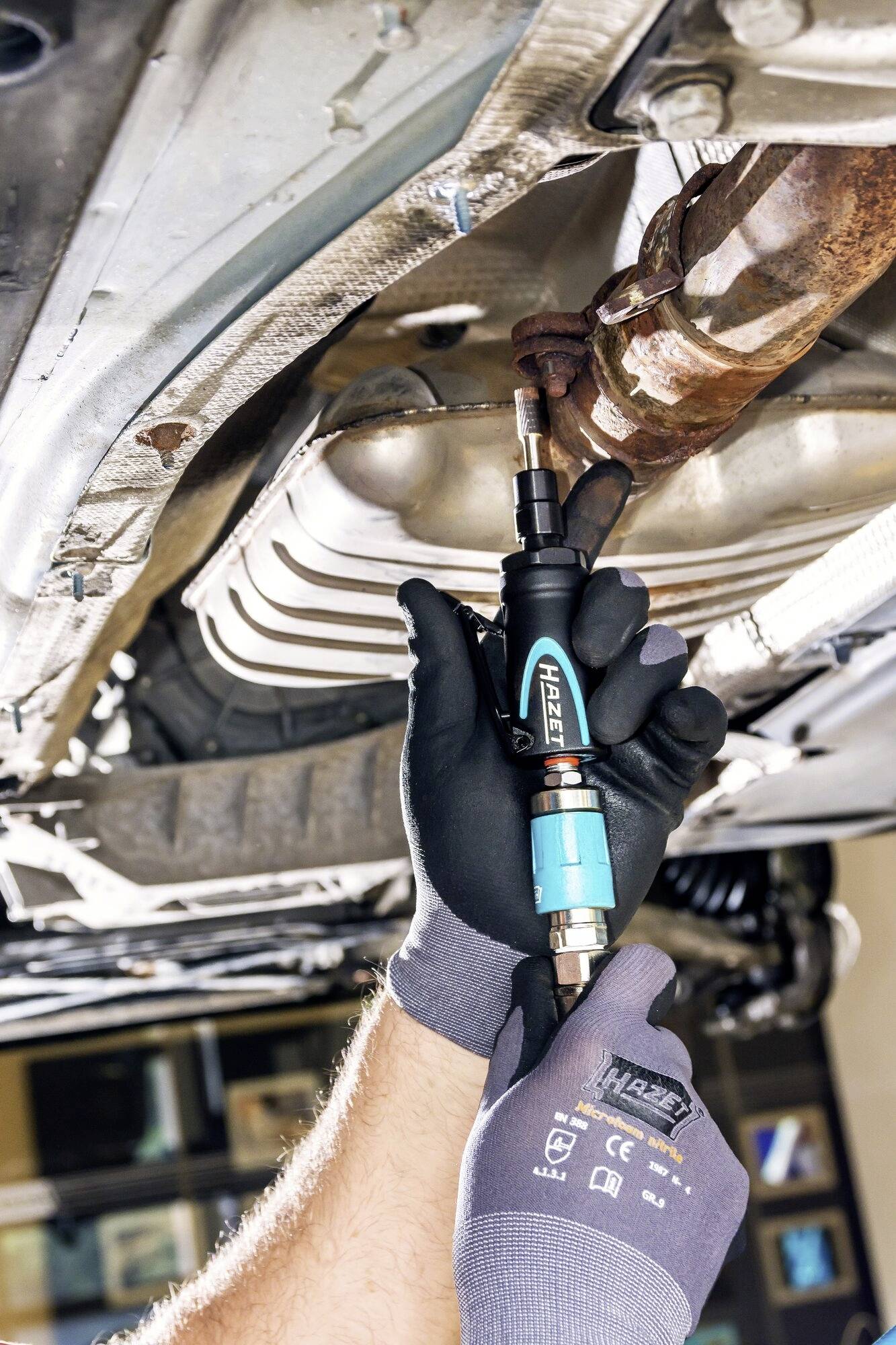 'Eine Person arbeitet unter einem Auto und verwendet ein Werkzeug, um eine Metallverbindung zu lösen.'