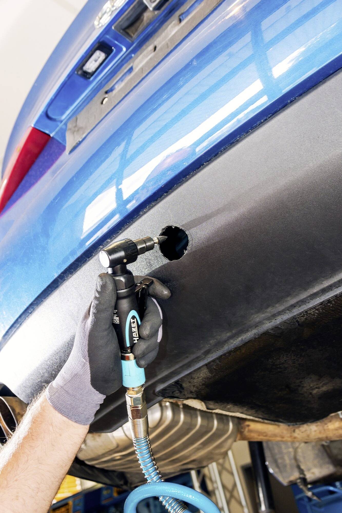 Person montiert eine Anhängerkupplung an ein blaues Auto in einer Werkstatt. Werkzeug und Autoauspuff sind sichtbar.