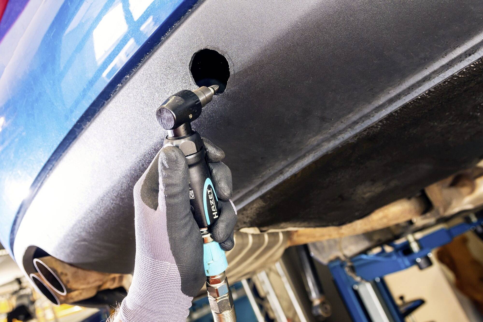 Ein Mechaniker repariert den Auspuff eines blauen Autos. Er verwendet ein spezielles Werkzeug, um ein Loch im Auspuff zu vergrößern.