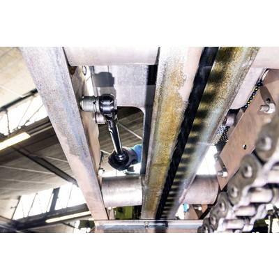Blick auf eine industrielle Maschine von unten, ein Arbeiter repariert sie mit einem Werkzeug. Sichtbare Ketten und Metallstrukturen.