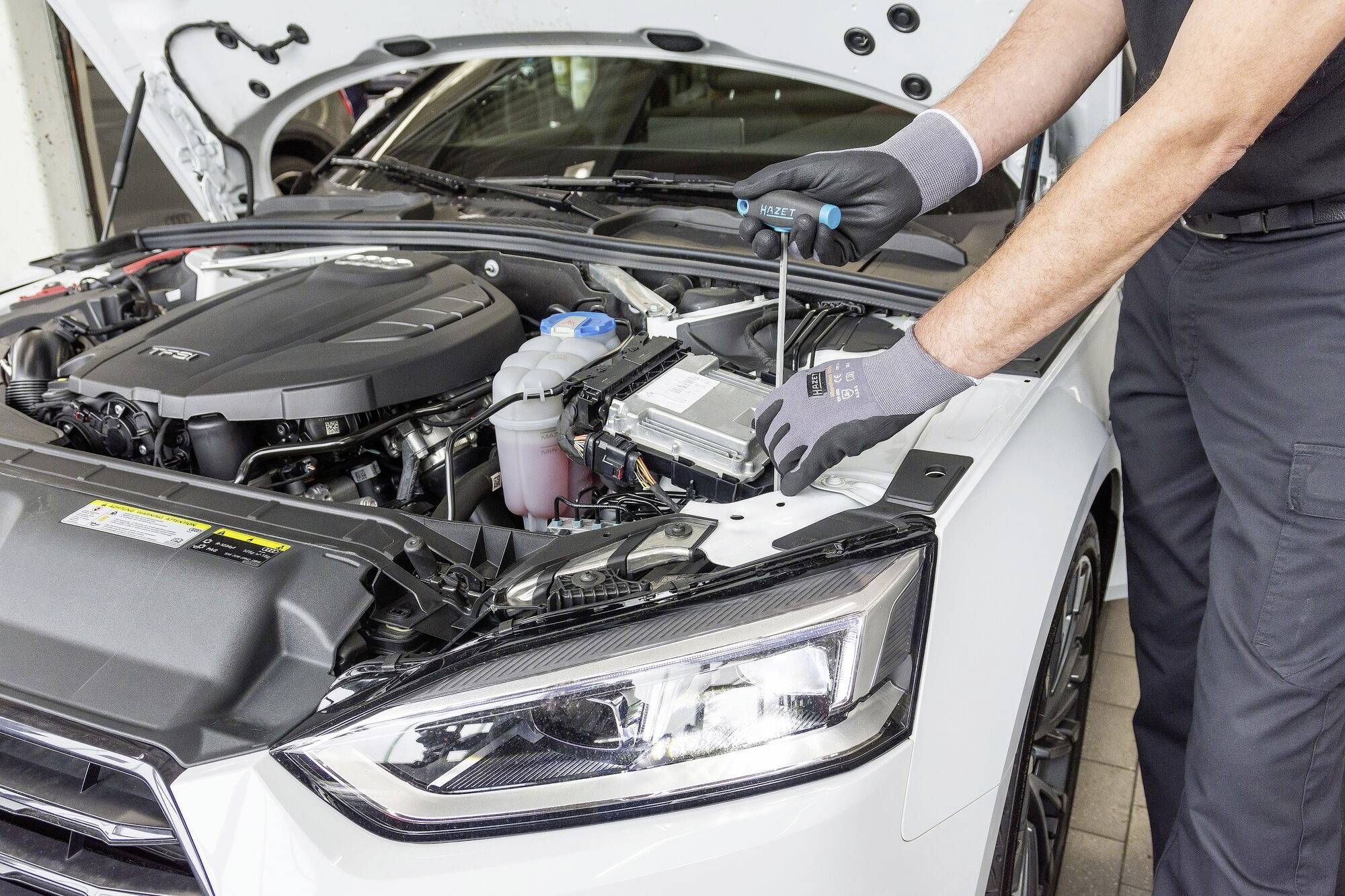 'Mechaniker arbeitet am Motor eines weißen Autos mit offenem Motorraum und benutzt einen Schraubendreher.'