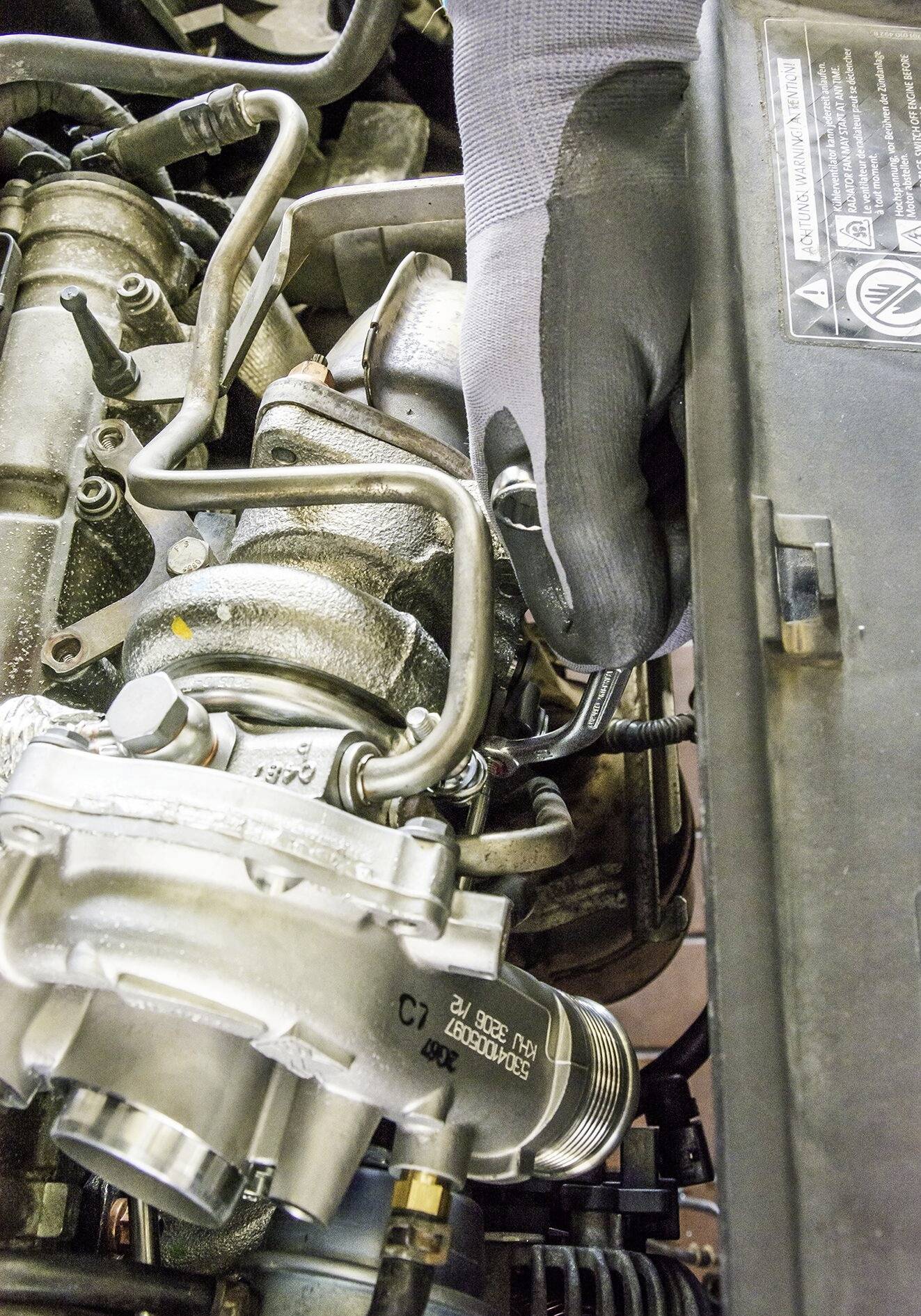 Ein Mechaniker repariert einen Automotor. Eine Hand im schwarzen Handschuh hält ein Werkzeug, um an einer Metallkomponente zu arbeiten.