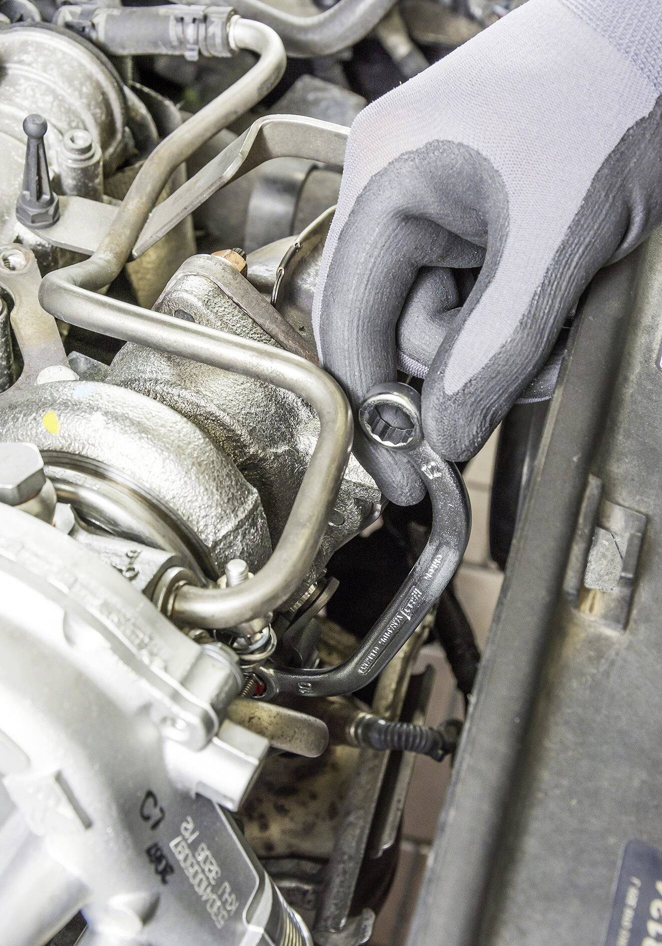 Eine Person mit grauem Handschuh zieht mit einem Schraubenschlüssel an einer Mutter im Motorraum eines Autos, um Reparaturen durchzuführen.