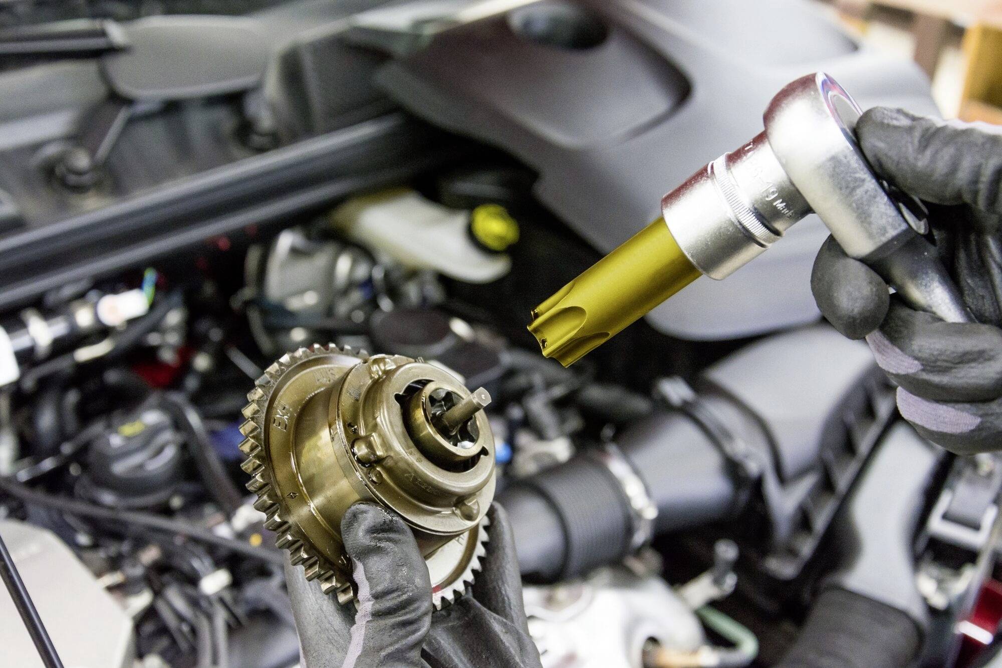 Ein Mechaniker hält einen Autoteil in der einen Hand und ein Spezialwerkzeug in der anderen vor einem geöffneten Motorraum.