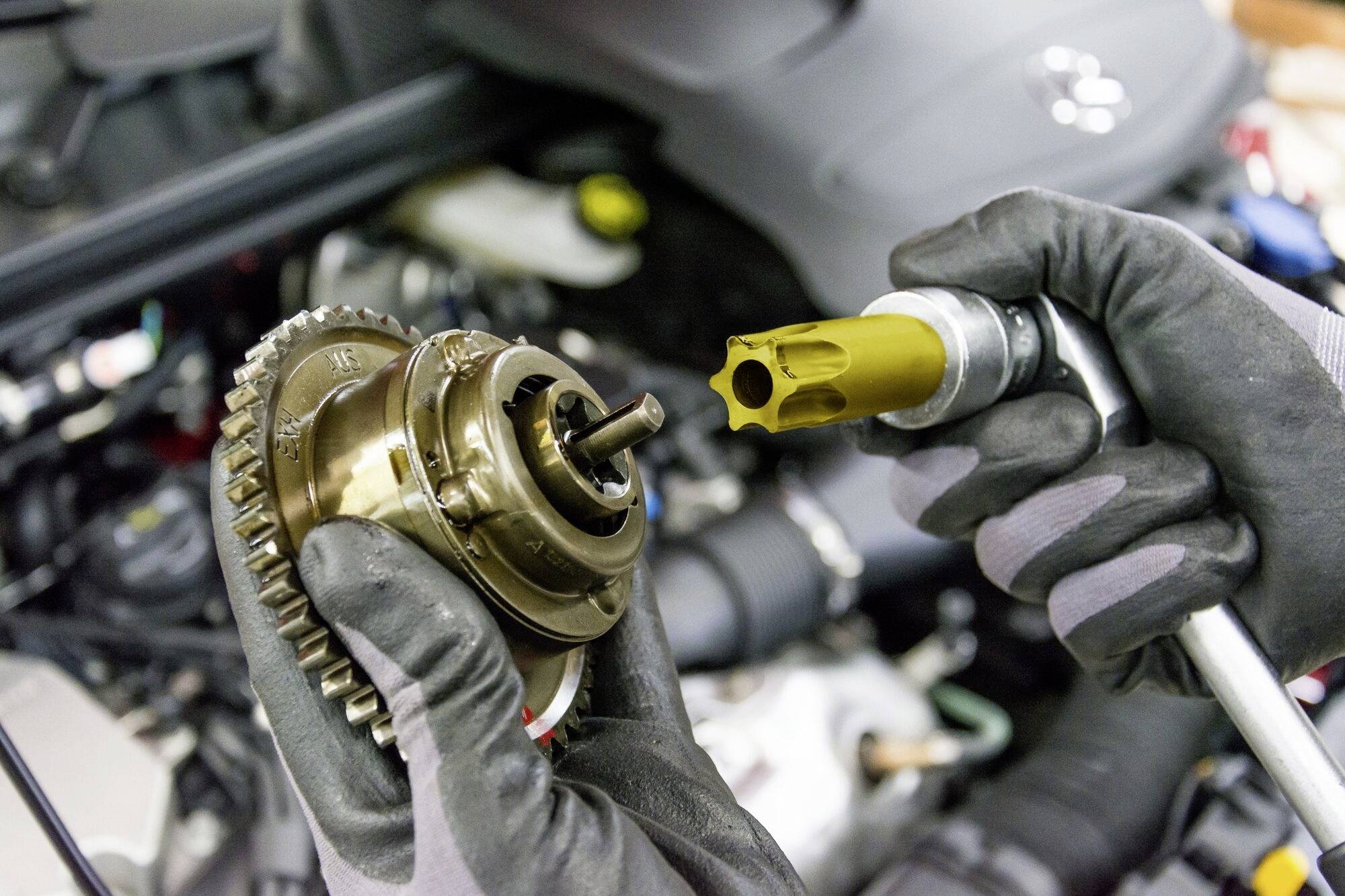 Mechaniker mit Handschuhen hält ein Autoteil und ein Werkzeug, während er an einem Motor arbeitet.