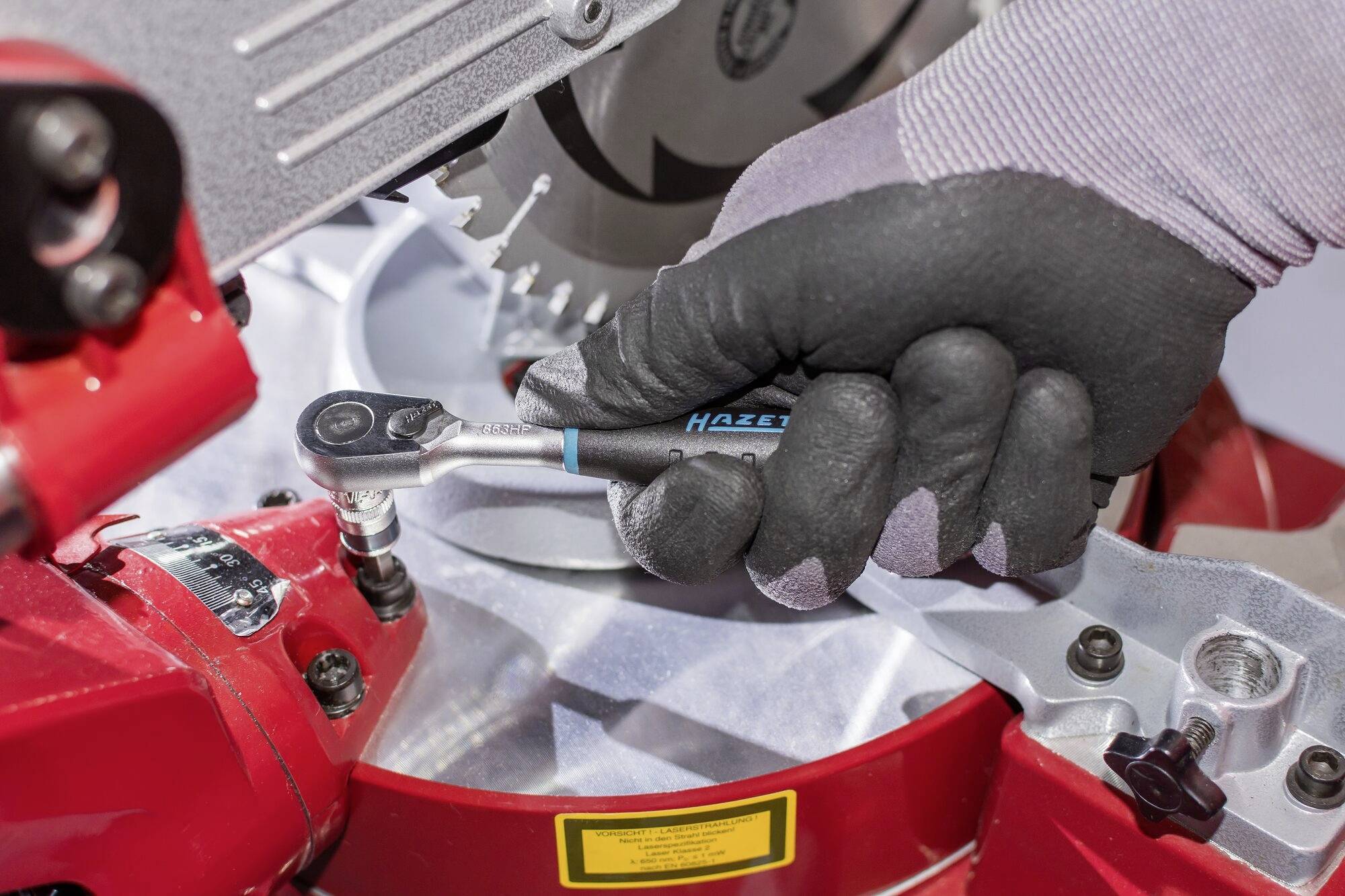 'Nahaufnahme einer Hand in einem Handschuh, die mit einem Schraubenschlüssel an einer roten Kreis- oder Tischsäge arbeitet.'