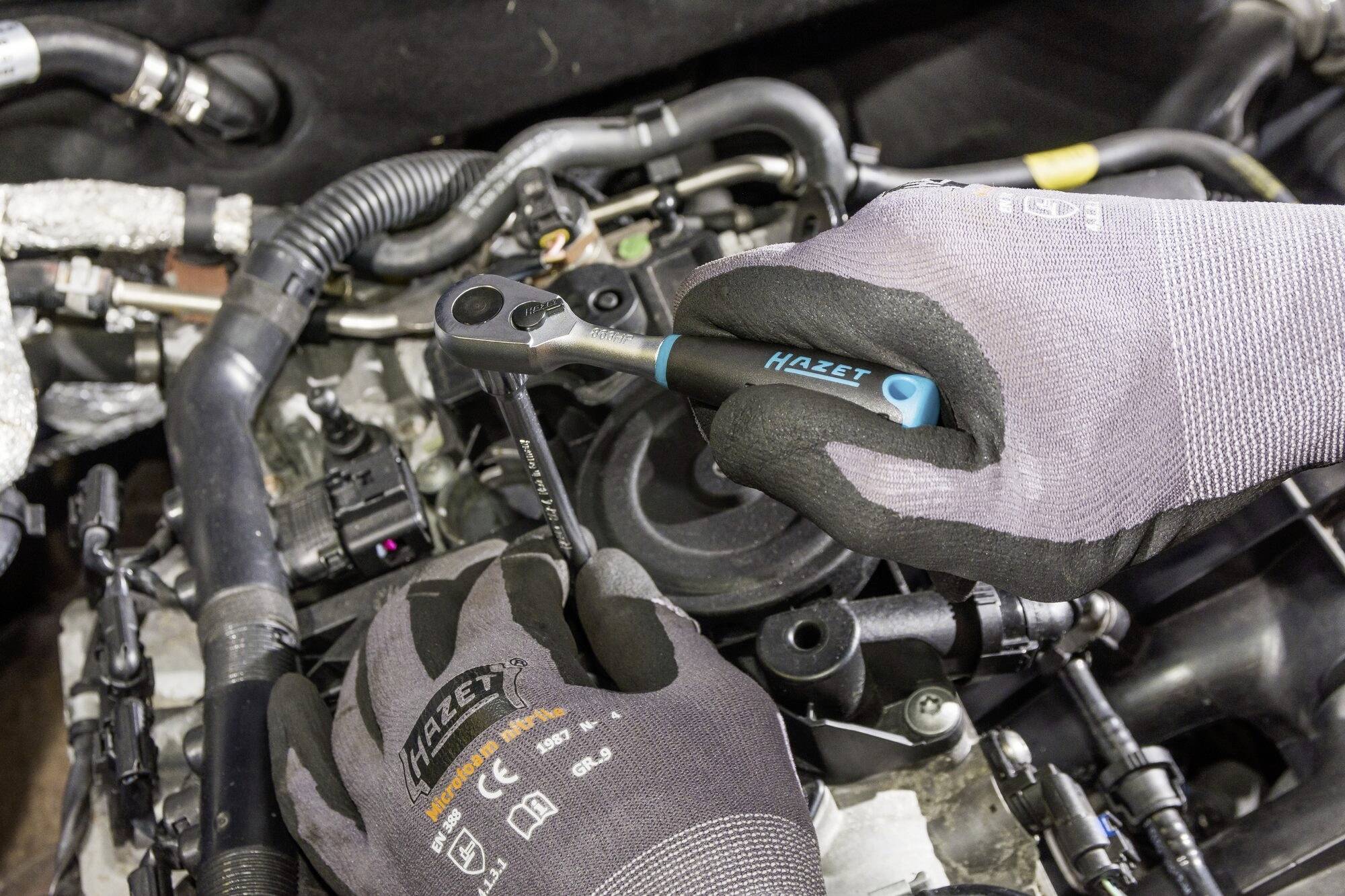 Eine Person repariert einen Motor mit einem Schraubenschlüssel. Die Hände tragen Schutzhandschuhe, im Hintergrund sind Motorenkomponenten sichtbar.