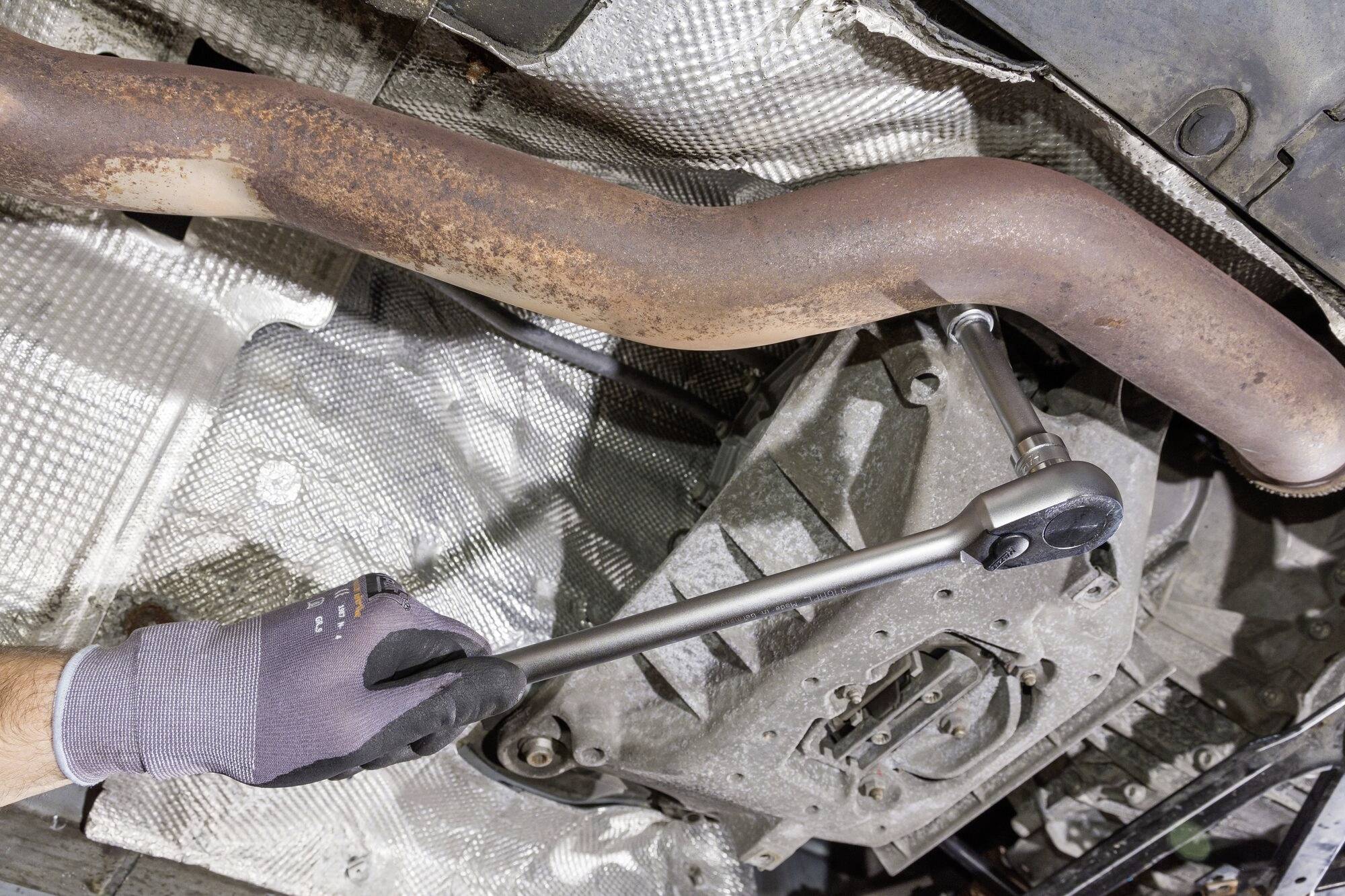 Mechanikerhand mit Handschuh benutzt Ratsche zum Lösen einer Schraube am Auspuff einer Fahrzeugunterseite.