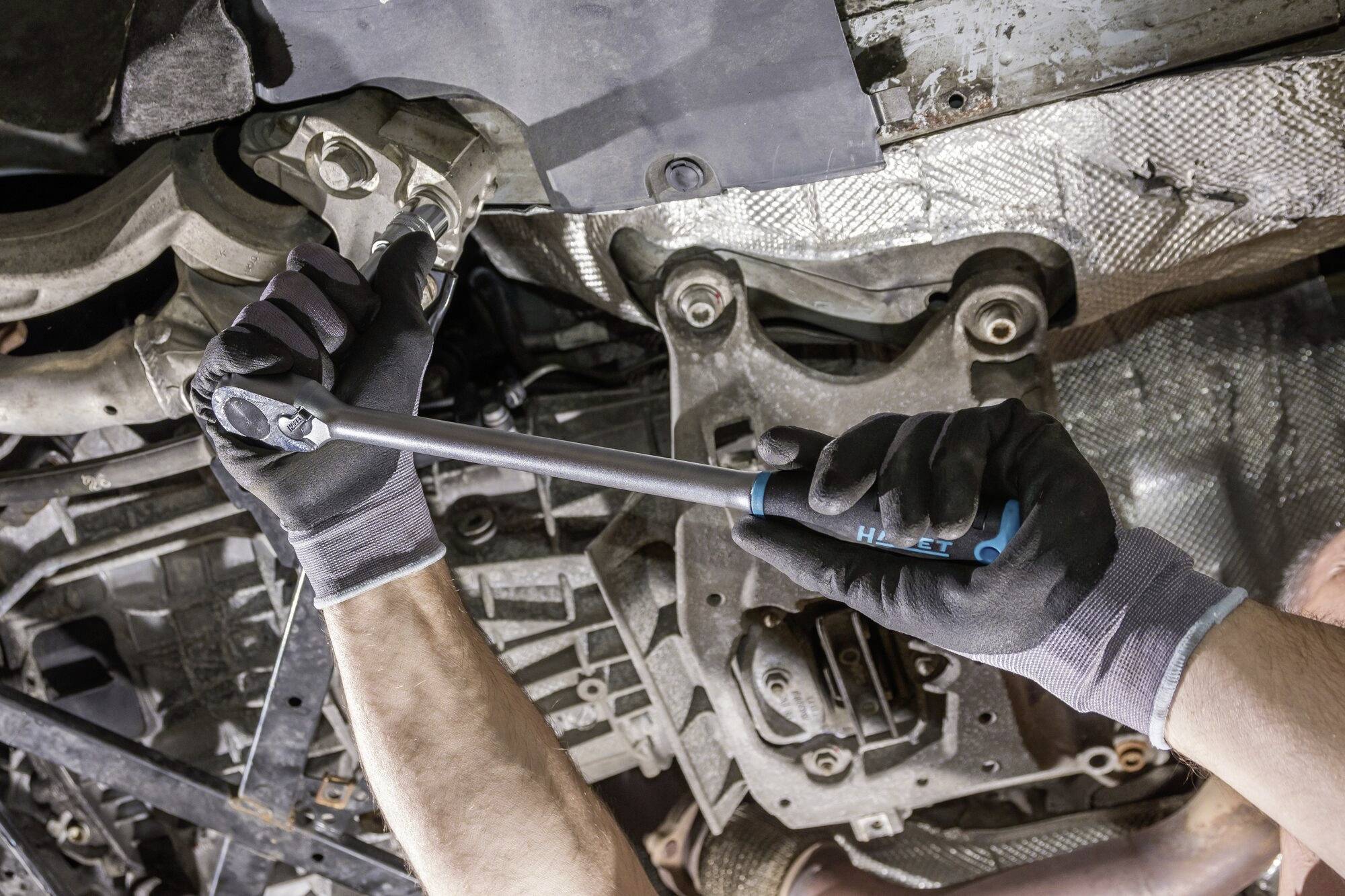 'Mechaniker zieht Schraube an Autochassis an, Nahaufnahme. Werkzeug mit Drehmomentschlüssel, Arbeitshandschuhe, Metallstruktur erkennbar.'