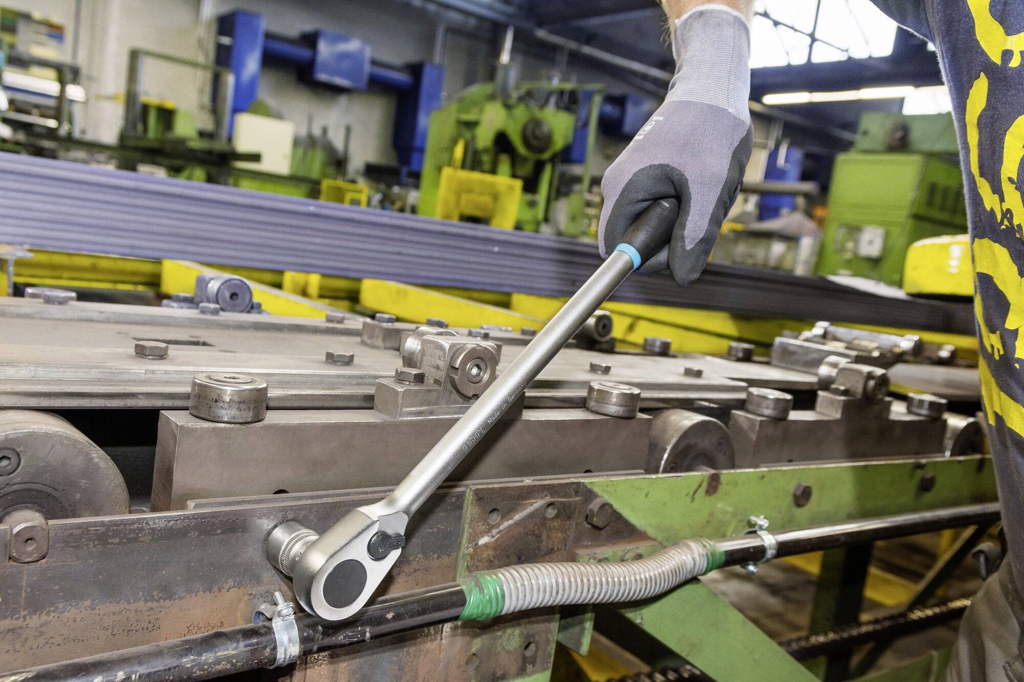 Ein Arbeiter verwendet einen Drehmomentschlüssel in einer Fabrikumgebung, um Maschinen zu warten. Im Hintergrund sind grüne Maschinen sichtbar.