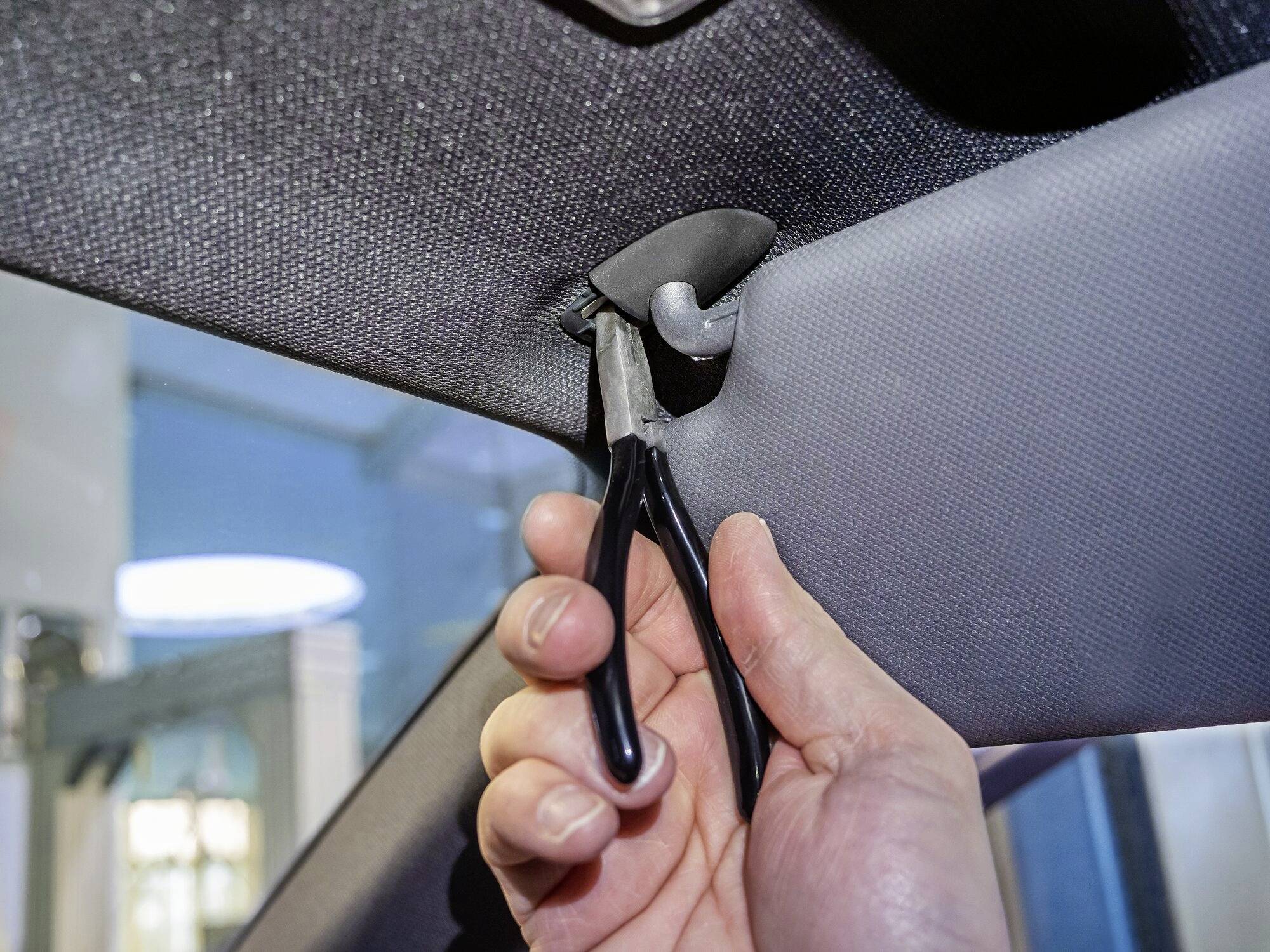 Eine Hand hält eine Zange und entfernt eine Halterung von der Autodecke.