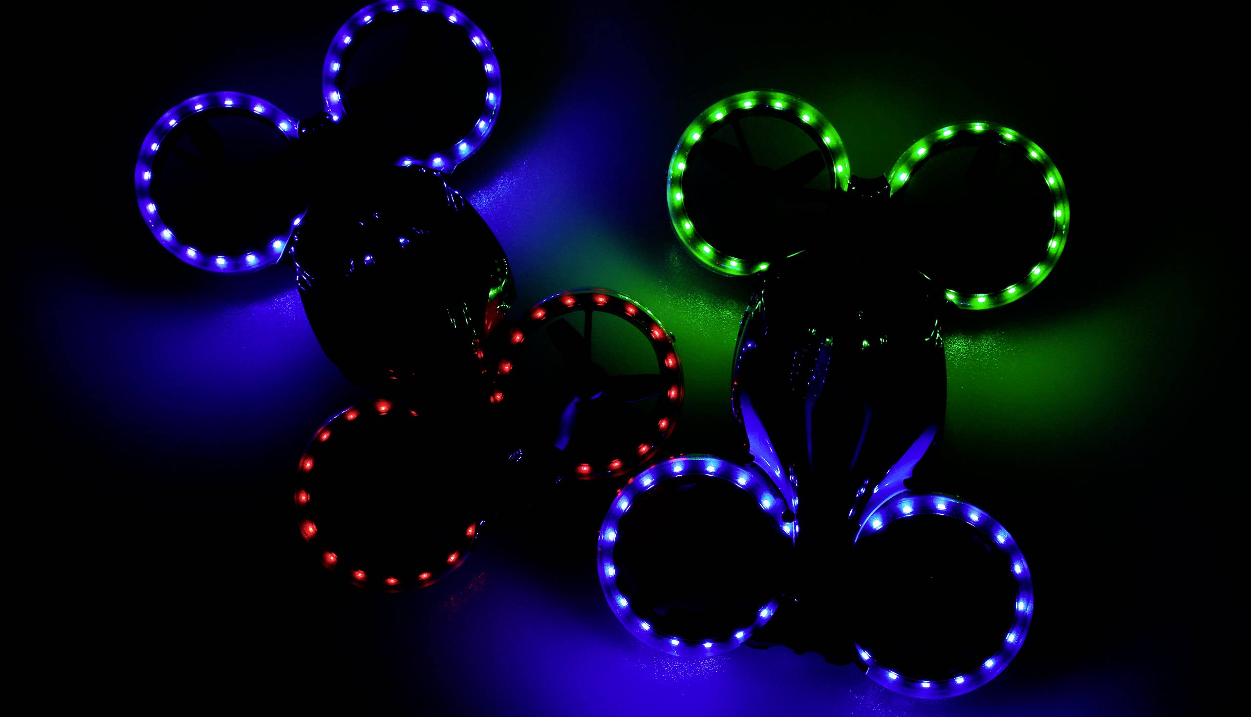 Zwei Drohnen mit bunten LED-Lichtern, die rot, blau und grün leuchten, fliegen in der Dunkelheit. Die Ringe um die Propeller sind beleuchtet.