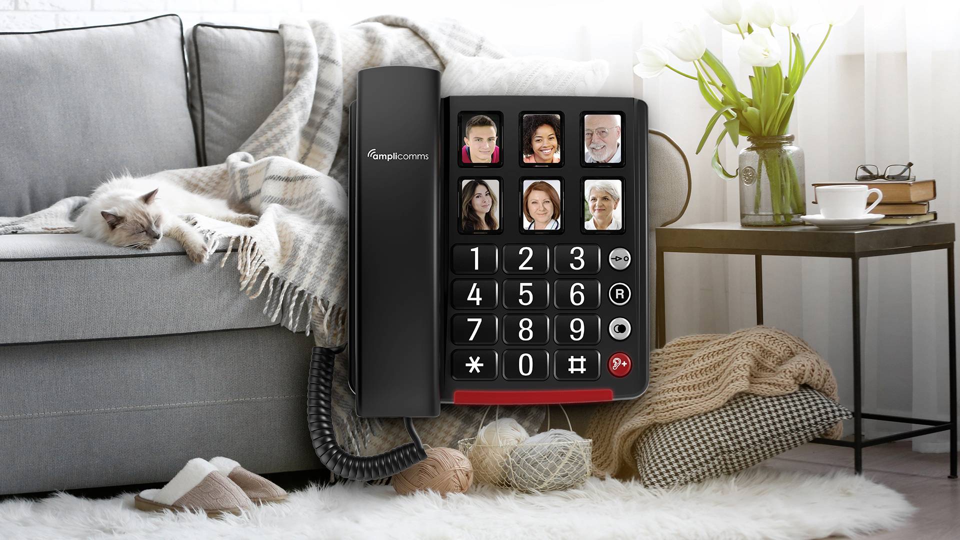 Ein Telefon mit großen Tasten und Fotos, um Kontakte zu speichern. Im Hintergrund schläft eine Katze auf einem Sofa neben einem Beistelltisch mit Blumen.
