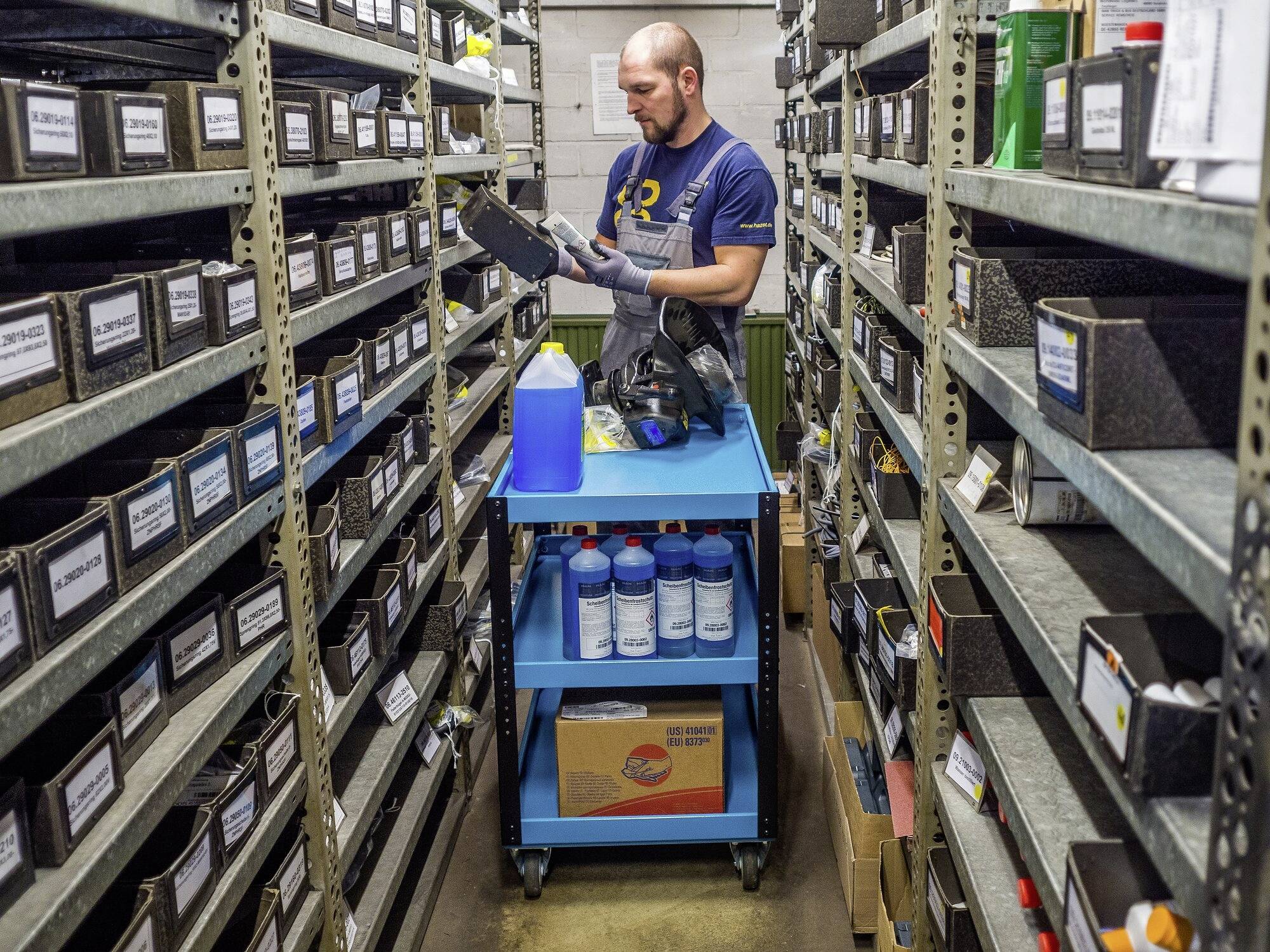Ein Mann sortiert Autoteile in einem Lagerraum. Er steht vor einem Regal mit beschrifteten Kartons und hält ein Teil in der Hand.