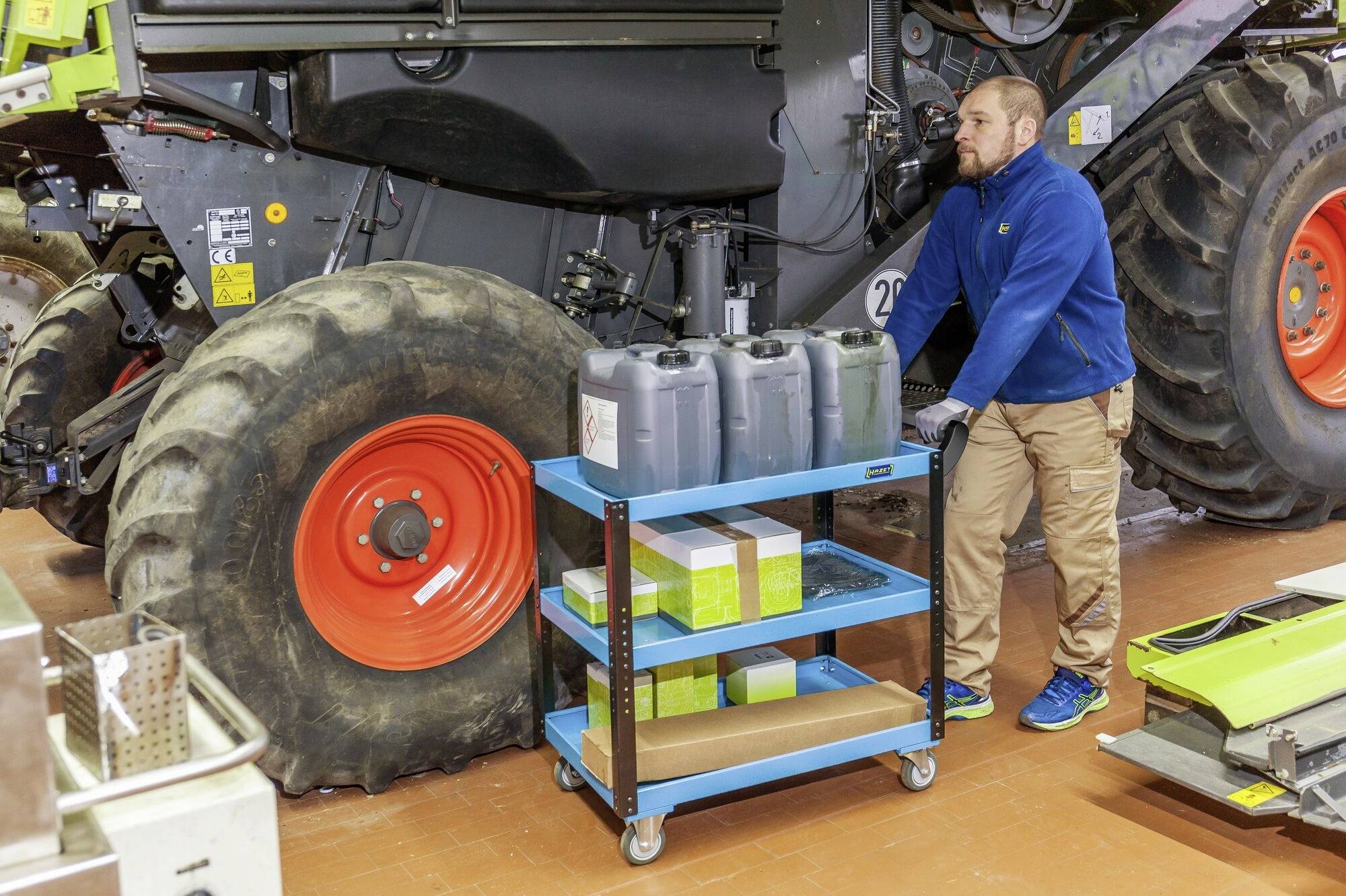 Ein Mann in blauer Arbeitskleidung schiebt einen blauen Wagen mit Kanistern und Boxen in einer Werkstatt neben einem großen Reifen.
