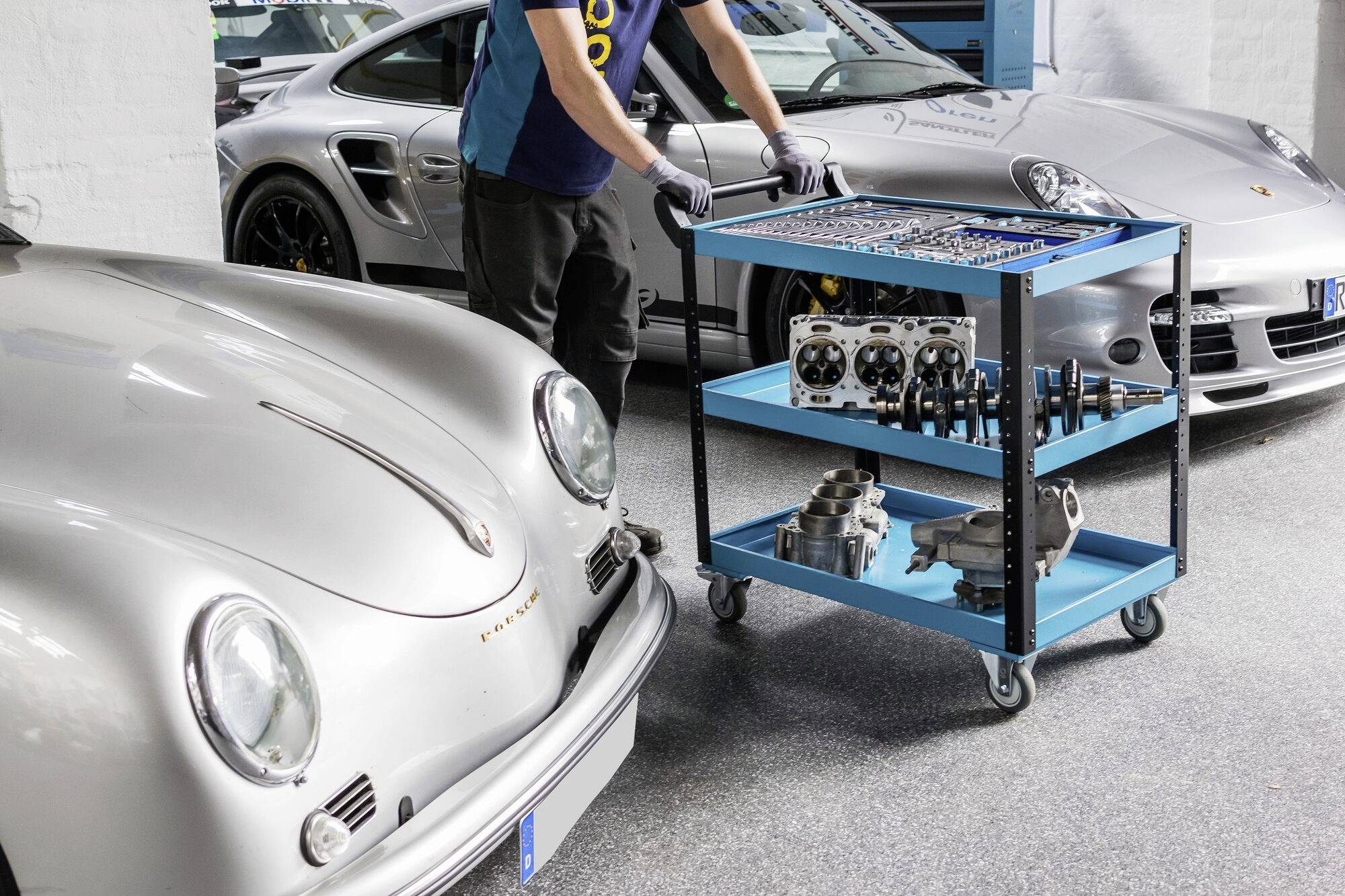 Eine Person arbeitet in einer Werkstatt an einem Werkzeugwagen. Zwei silberne Sportwagen stehen dahinter geparkt.