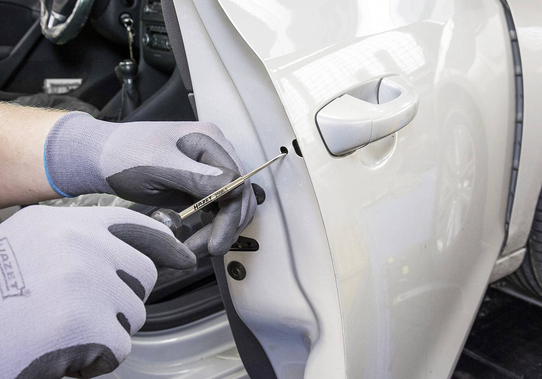 'Eine Person mit grauen Handschuhen verwendet einen Schraubendreher an der Tür eines weißen Autos. Es sieht nach einer Reparatur oder Wartung aus.'