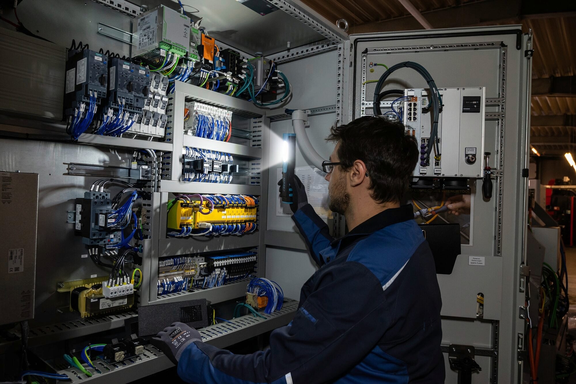 Ein Techniker arbeitet mit einer Taschenlampe an einem Schaltschrank, untersucht Verkabelungen und Komponenten in einem Industrieumfeld.