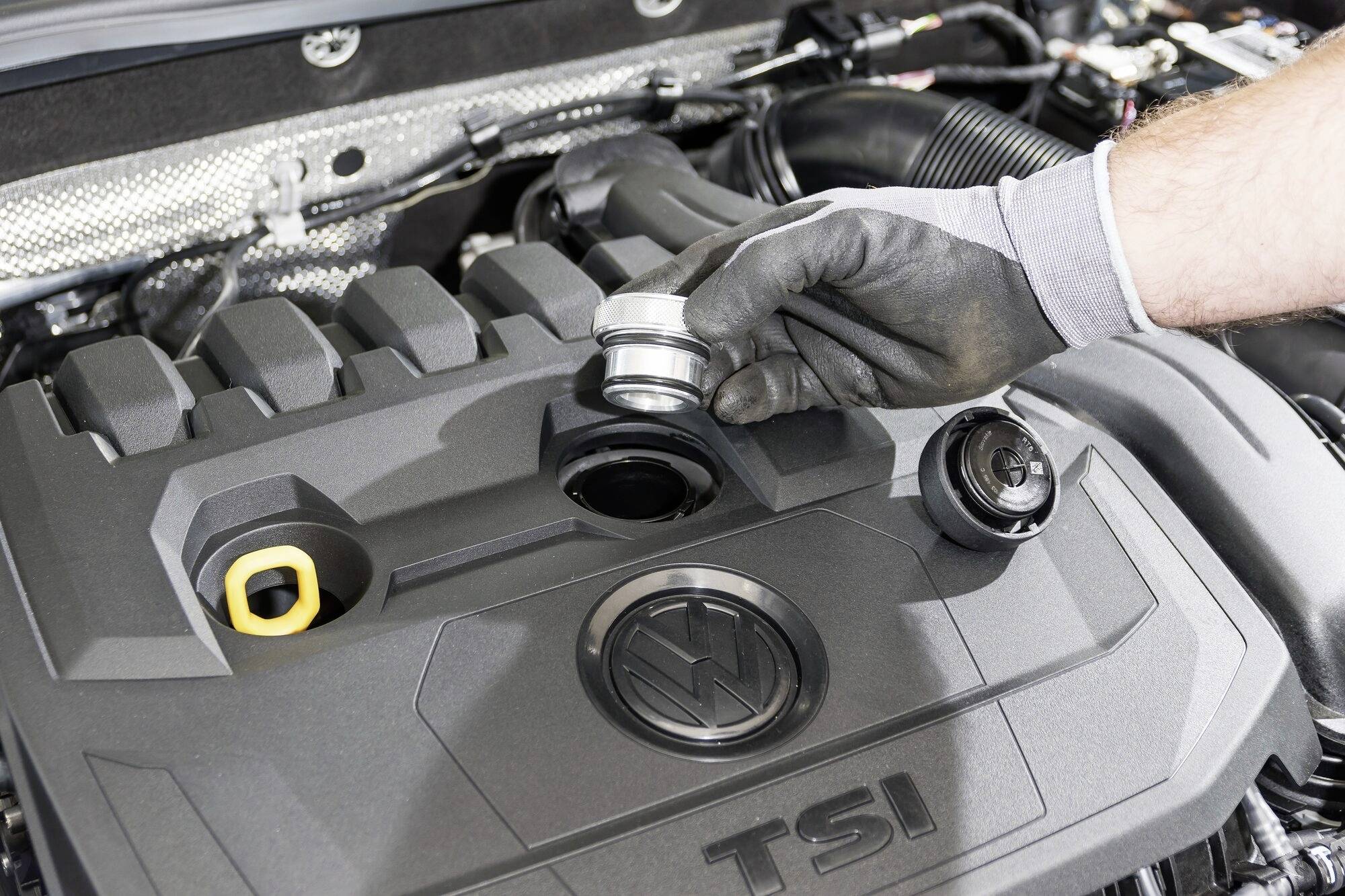 Eine behandschuhte Hand wechselt den Ölverschluss an einem TSI-Motor eines Autos. Der Motorhaubenbereich ist im Hintergrund sichtbar.