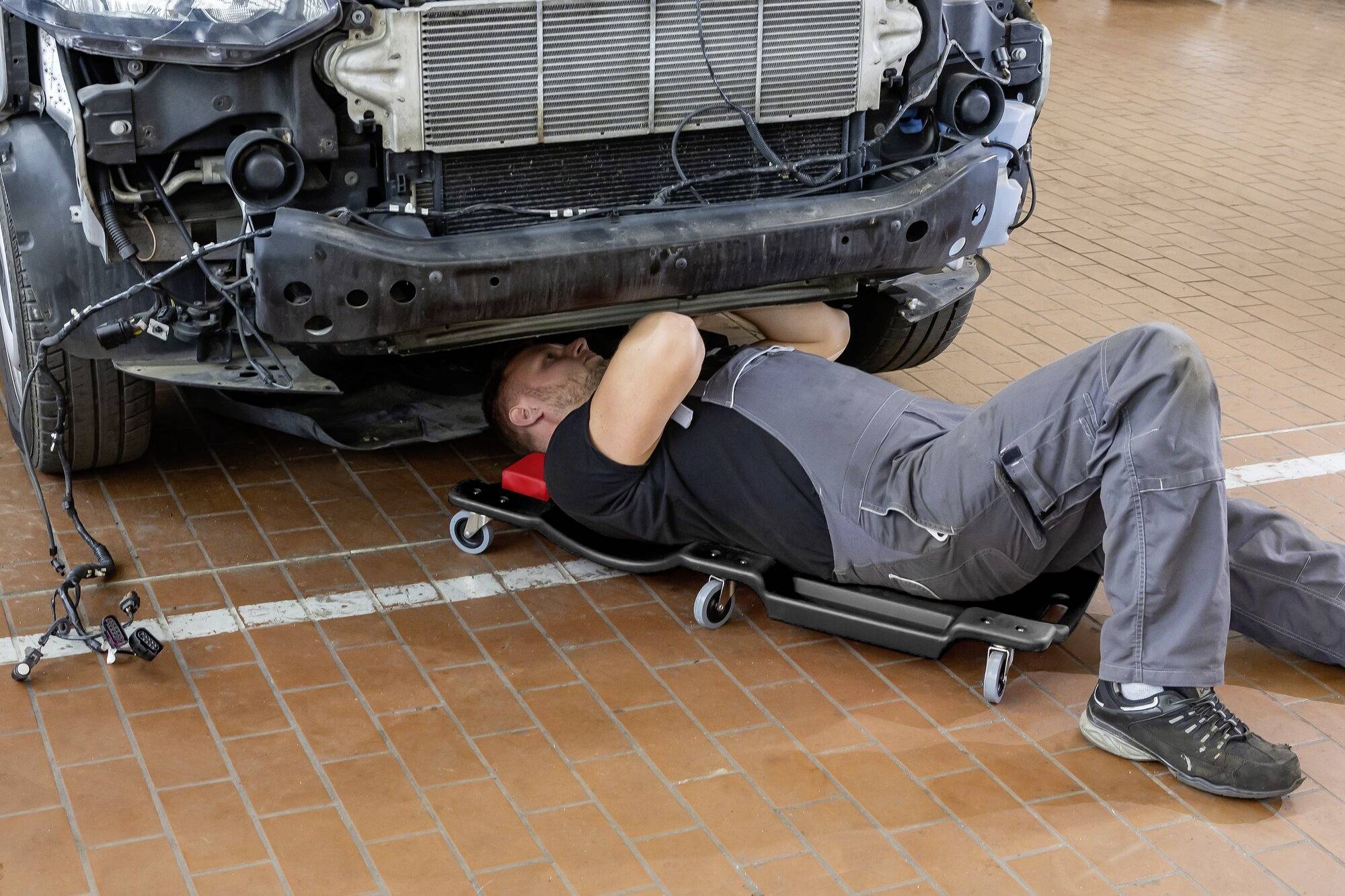 Ein Mechaniker arbeitet unter einem Auto, das auf einer Reparaturstation steht. Er liegt auf einem Rollbrett und trägt Arbeitskleidung.