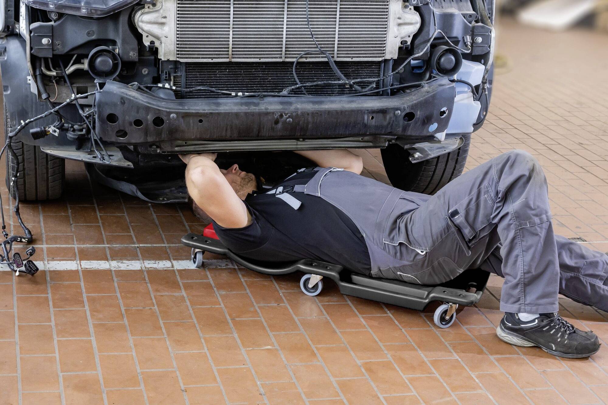 Eine Person liegt auf einem Rollbrett unter einem Auto und arbeitet am Fahrzeugunterboden in einer Werkstatt.