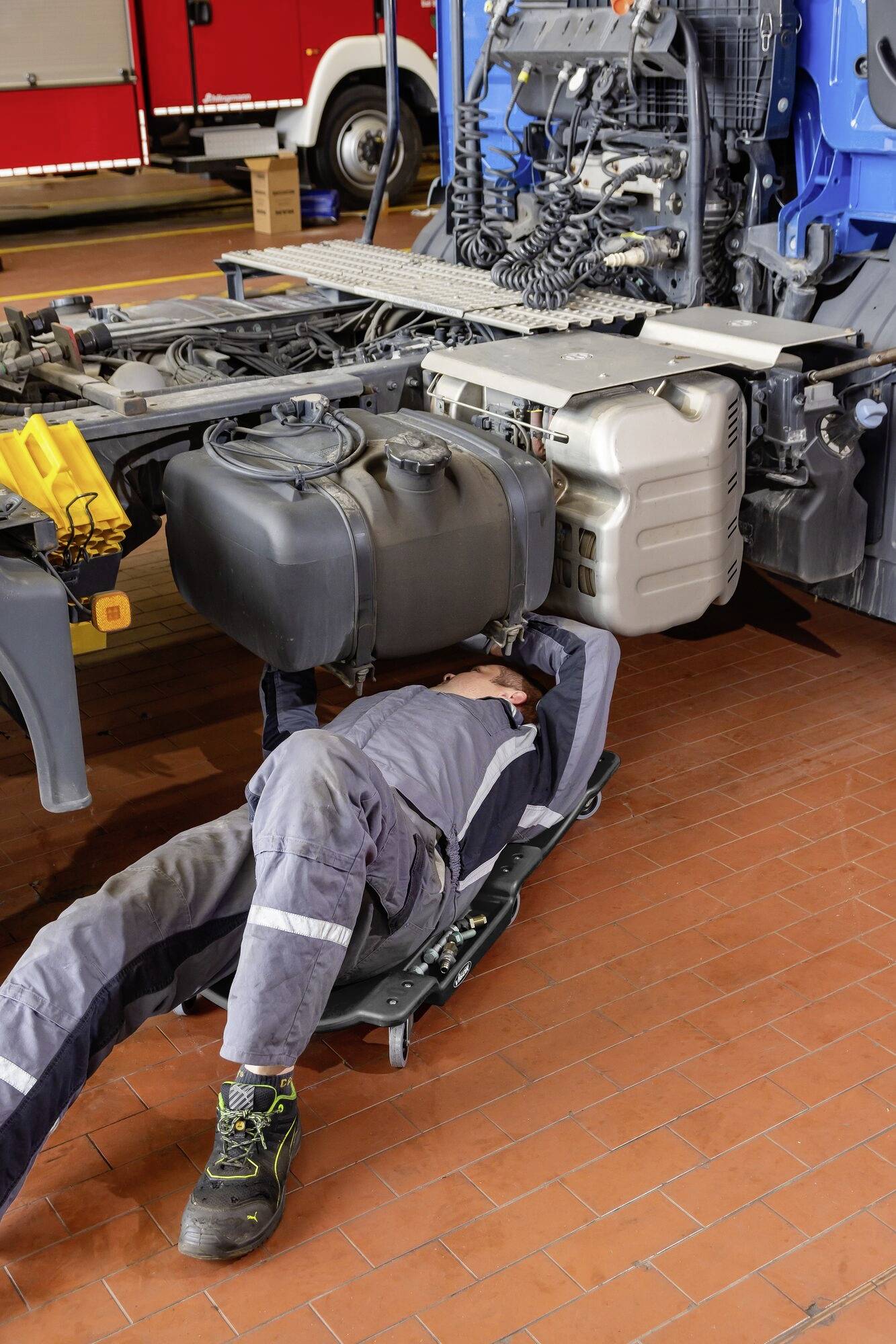 Eine Person in Arbeitskleidung repariert unter einem Lastwagen in einer Werkstatt. Im Hintergrund sind weitere Lkw zu sehen.