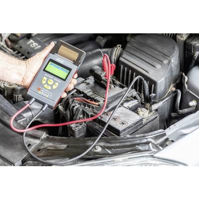 Eine Person hält ein Batterietestgerät am offenen Fahrzeugmotorraum. Das Gerät zeigt Informationen über den Batteriestatus an.